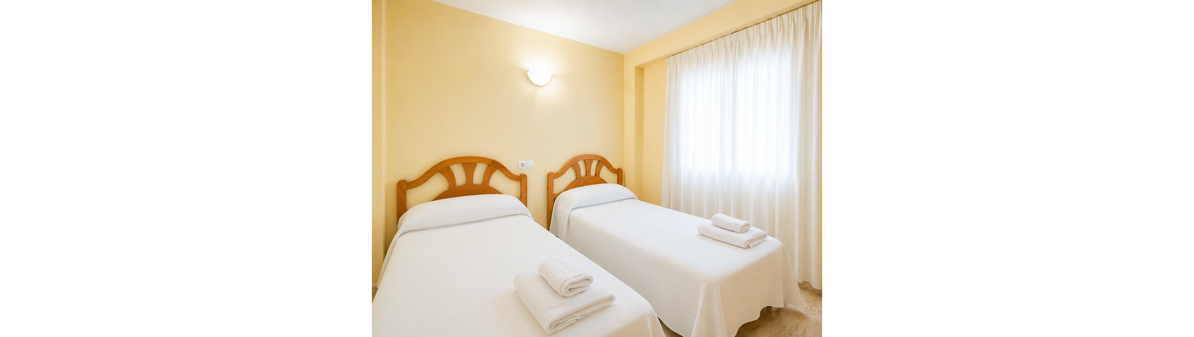 Simple twin beds with wooden headboards are neatly made with white linens and folded towels in a bright, minimalist room. Soft yellow walls and a wall-mounted light create a warm, inviting atmosphere. Sheer white curtains cover a large window, allowing diffused natural light to fill the space. The overall look is clean, orderly, and suitable for a hotel or guest accommodation.