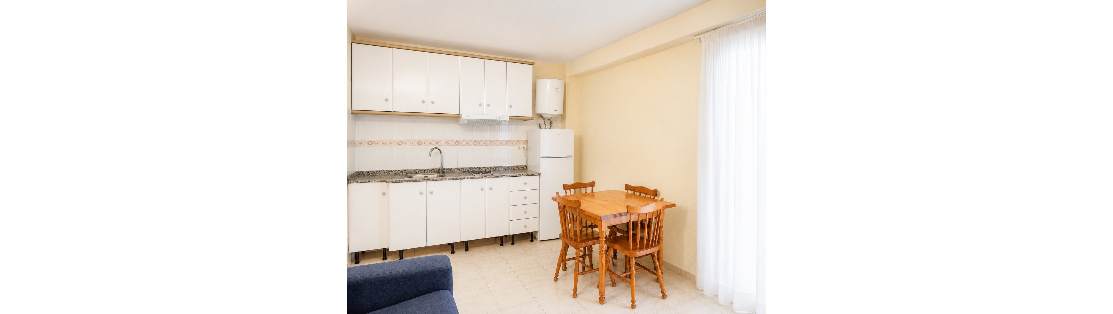 A small, bright kitchen features white cabinets and a granite countertop with a single metal faucet. A white refrigerator and water heater stand against the wall near the cooking area. A wooden table with four matching chairs occupies the center of the room, adding warmth to the neutral space. Light-colored tiled flooring and sheer curtains over a large window enhance the airy, minimalist feel.