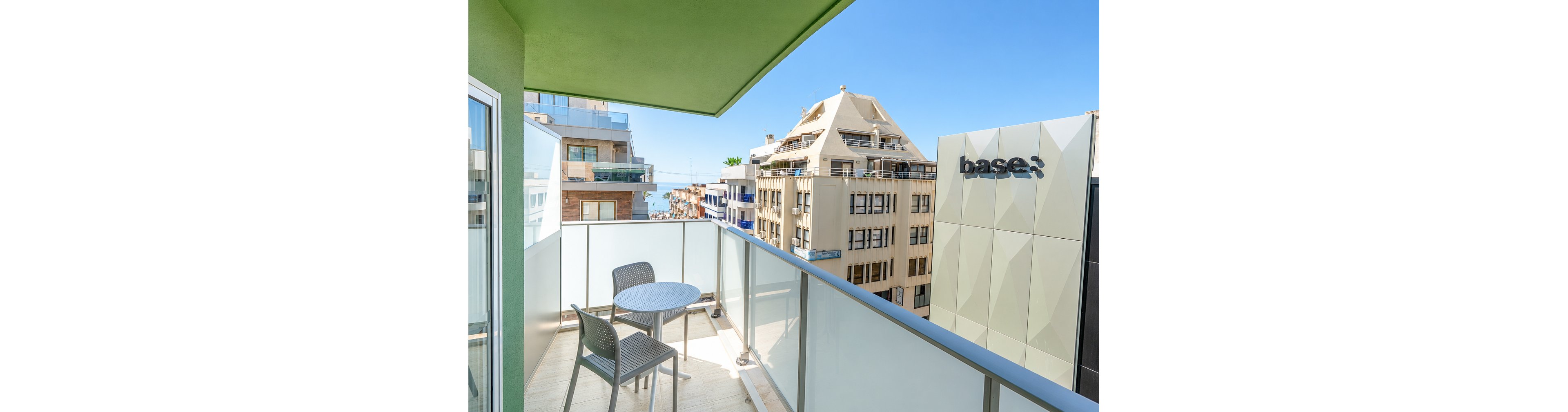 A small modern balcony features a round table and two chairs overlooking an urban streetscape. Glass railings frame the view of contemporary apartment buildings and a nearby structure with the clearly visible text "base:" on its facade. In the distance, a glimpse of the sea appears under a clear blue sky, suggesting a coastal city location. Bright daylight and clean architectural lines create a fresh, minimalist atmosphere.