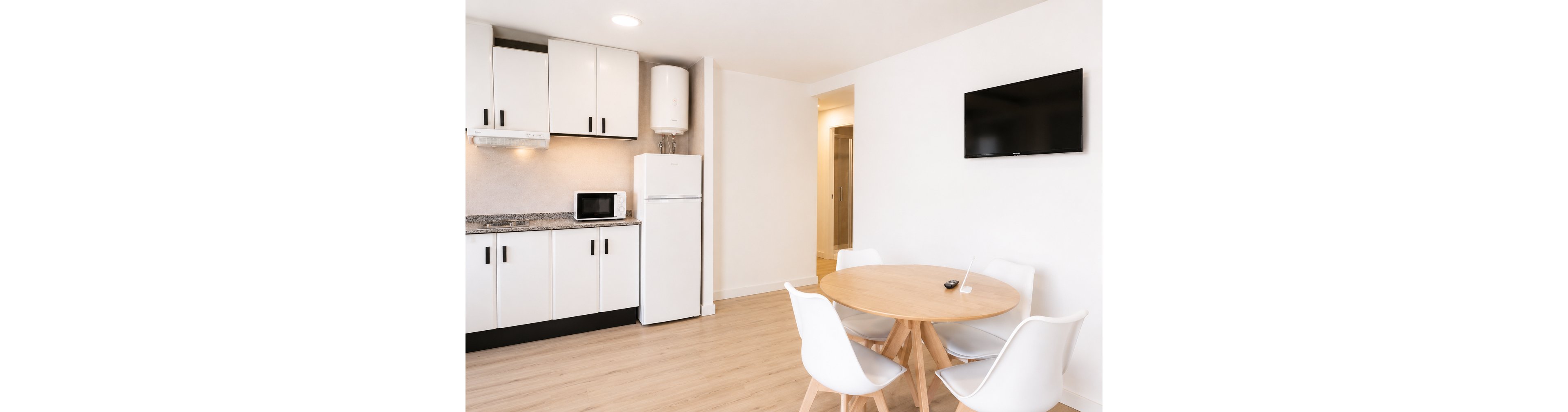 A compact modern kitchen features white cabinets, a refrigerator, microwave, and wall-mounted water heater along one side of the room. Adjacent to the kitchen, a round wooden dining table is surrounded by four white contemporary chairs on light wood flooring. A flat-screen television is mounted on the wall opposite the table, adding entertainment to the clean, functional space. The overall design is minimalist with a neutral palette emphasizing white surfaces and natural wood tones.