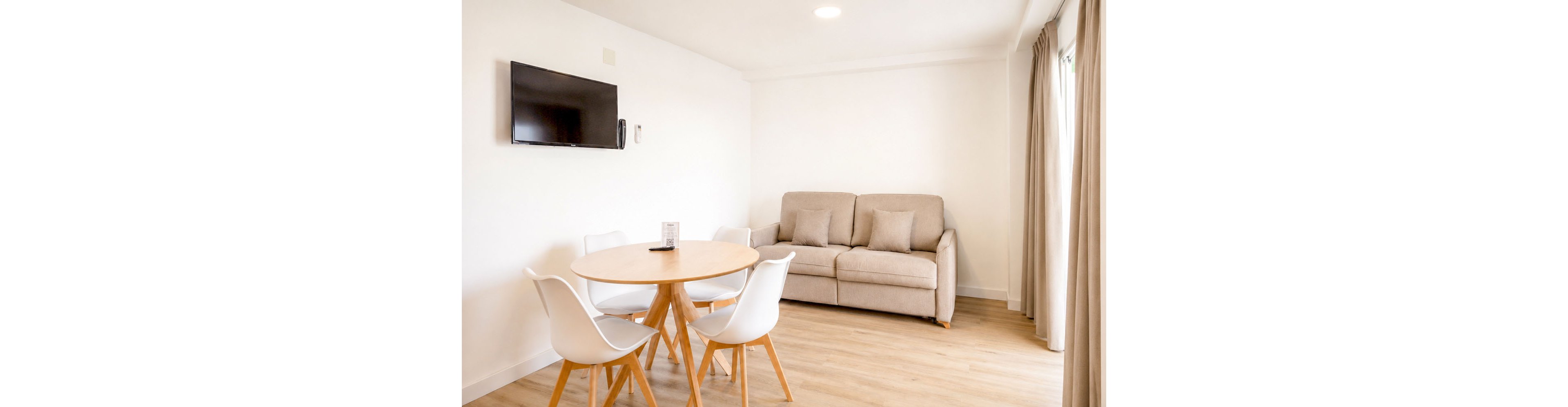 A modern living room features a beige sofa with matching cushions against a white wall. A round wooden dining table with four white chairs sits nearby on light wood flooring. A flat-screen television is mounted on the wall, with a small device or intercom beside it. Floor-to-ceiling curtains frame a window, allowing natural light to brighten the clean, minimalist space.