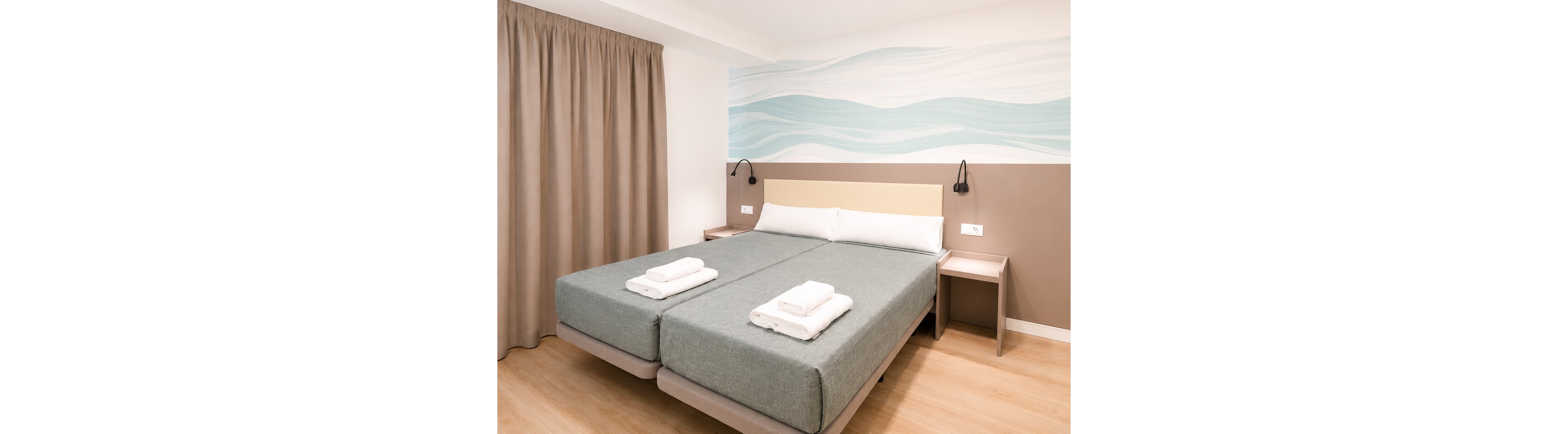 Bright, minimalist hotel room featuring two twin beds pushed together with neatly folded white towels on top. The space includes light wood flooring, beige curtains, and simple bedside tables with wall-mounted reading lamps. A soft blue and white wave mural decorates the wall above the headboard, adding a calm, coastal touch. The overall atmosphere is clean, modern, and inviting, suitable for comfortable short stays.