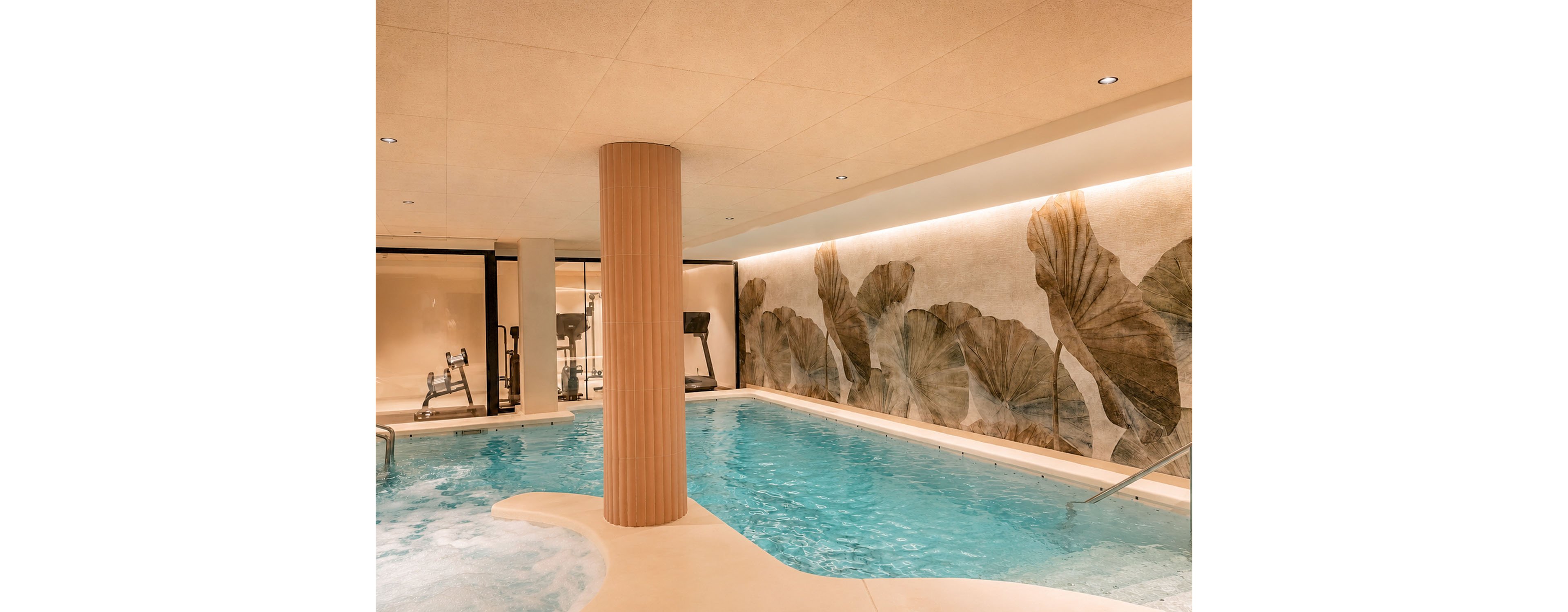 An indoor spa-style swimming pool is shown with gently rippling turquoise water and a curved entry area. A textured column rises from the pool deck, and a large botanical mural decorates the wall along one side. Warm ambient lighting runs above the mural, creating a relaxing atmosphere. In the background, behind glass, gym and fitness equipment are visible, suggesting a combined wellness and exercise space.