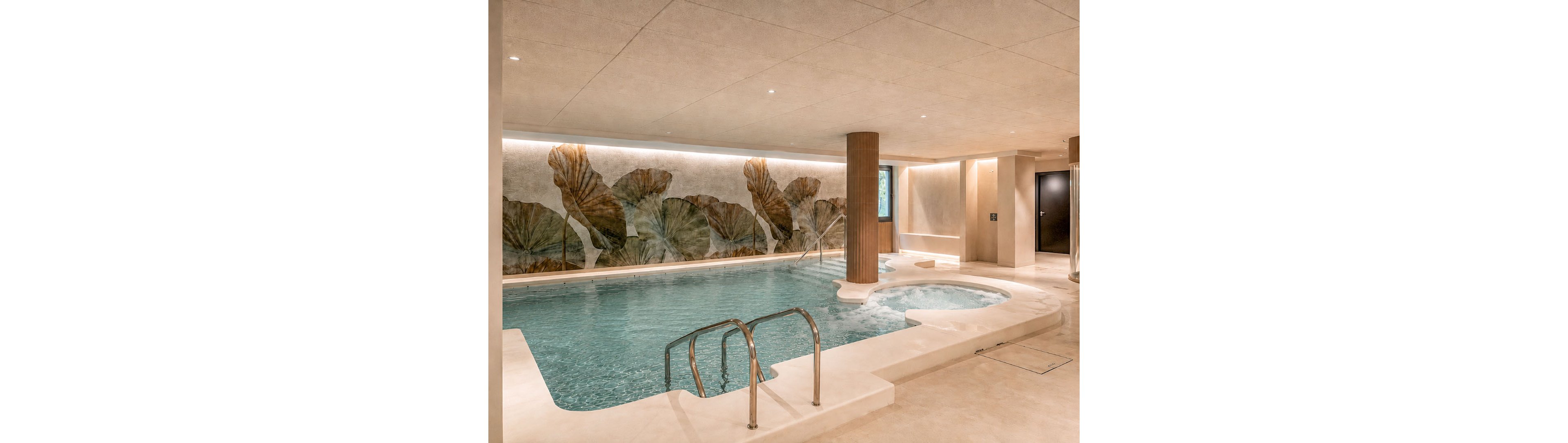 An elegant indoor spa pool features a curved main basin and a smaller circular whirlpool area. The space is finished in light neutral stone with recessed ceiling lights creating a calm, inviting atmosphere. A large wall mural of oversized leaves adds an artistic focal point behind the water. Stainless steel handrails, a central column, and minimal doors complete the contemporary spa design.