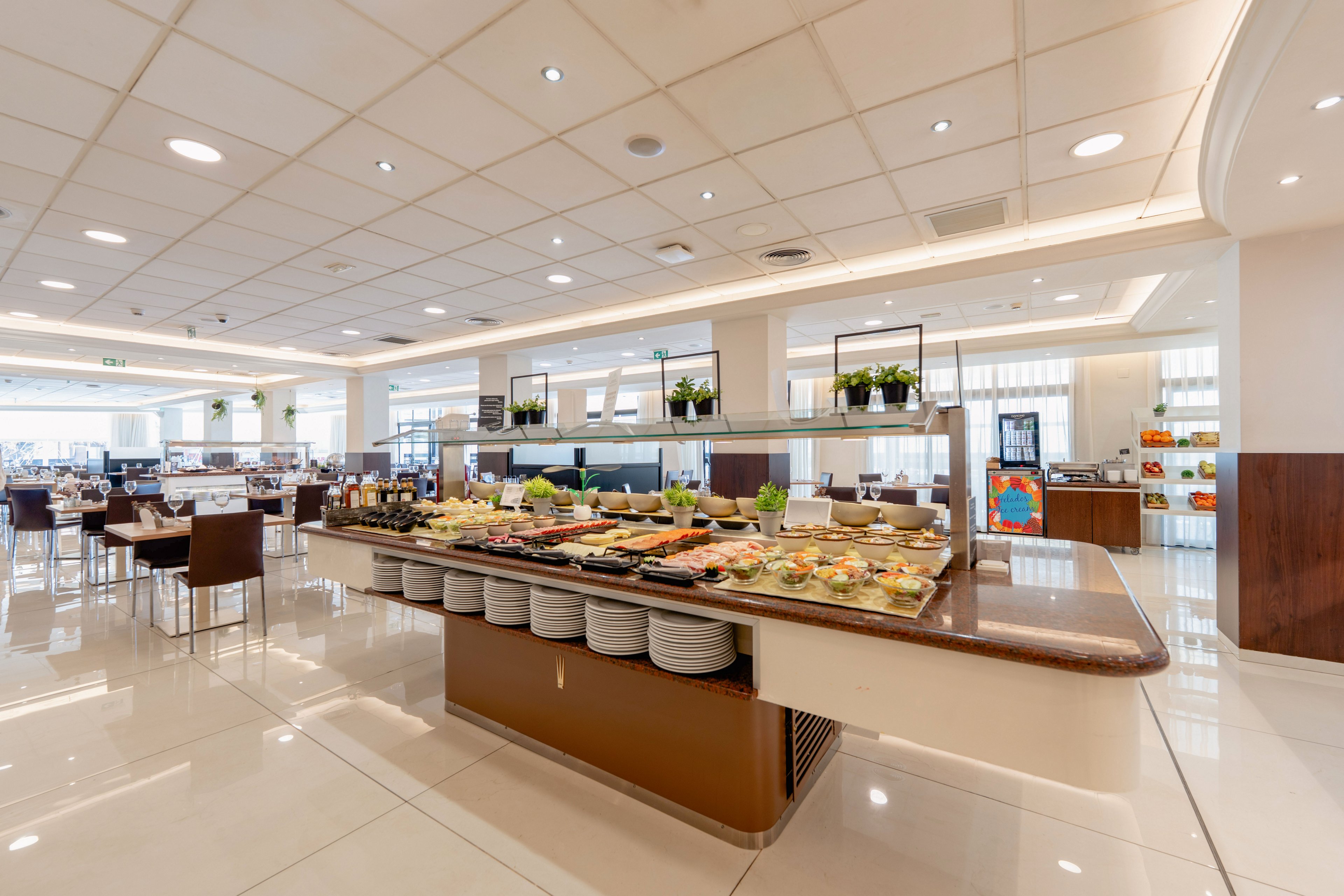A spacious, contemporary restaurant features a central buffet island filled with assorted salads, fruits, and cold dishes. Stacks of white plates are neatly arranged beneath the counter, ready for guests. The bright interior has glossy tiled floors, recessed ceiling lights, and rows of tables and chairs in the background. Potted plants and organized serving stations contribute to a clean, inviting dining atmosphere.