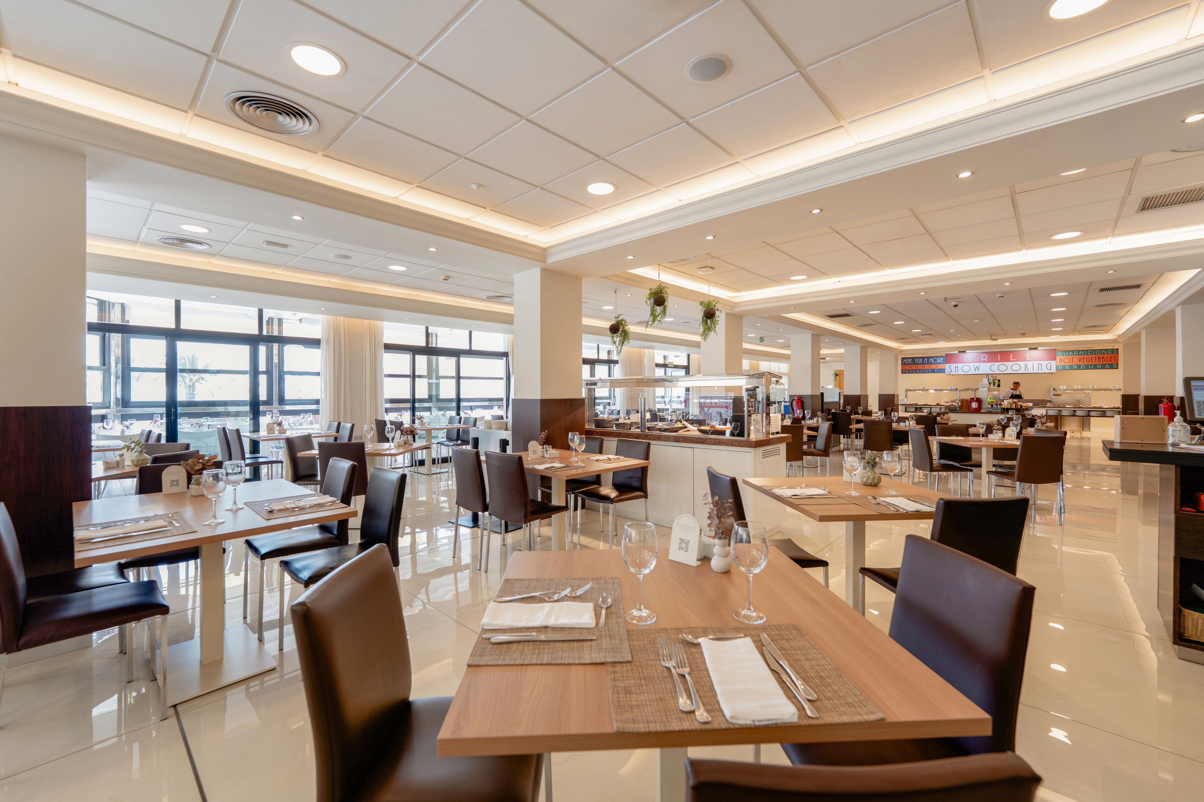 Spacious contemporary restaurant dining room with neatly arranged wooden tables and brown chairs. Place settings with cutlery, napkins, and glassware are prepared, suggesting readiness for guests. Large windows allow abundant natural light, highlighting the glossy tiled floor and clean ceiling design. The background shows a buffet or service counter area, creating a welcoming, casual dining atmosphere.