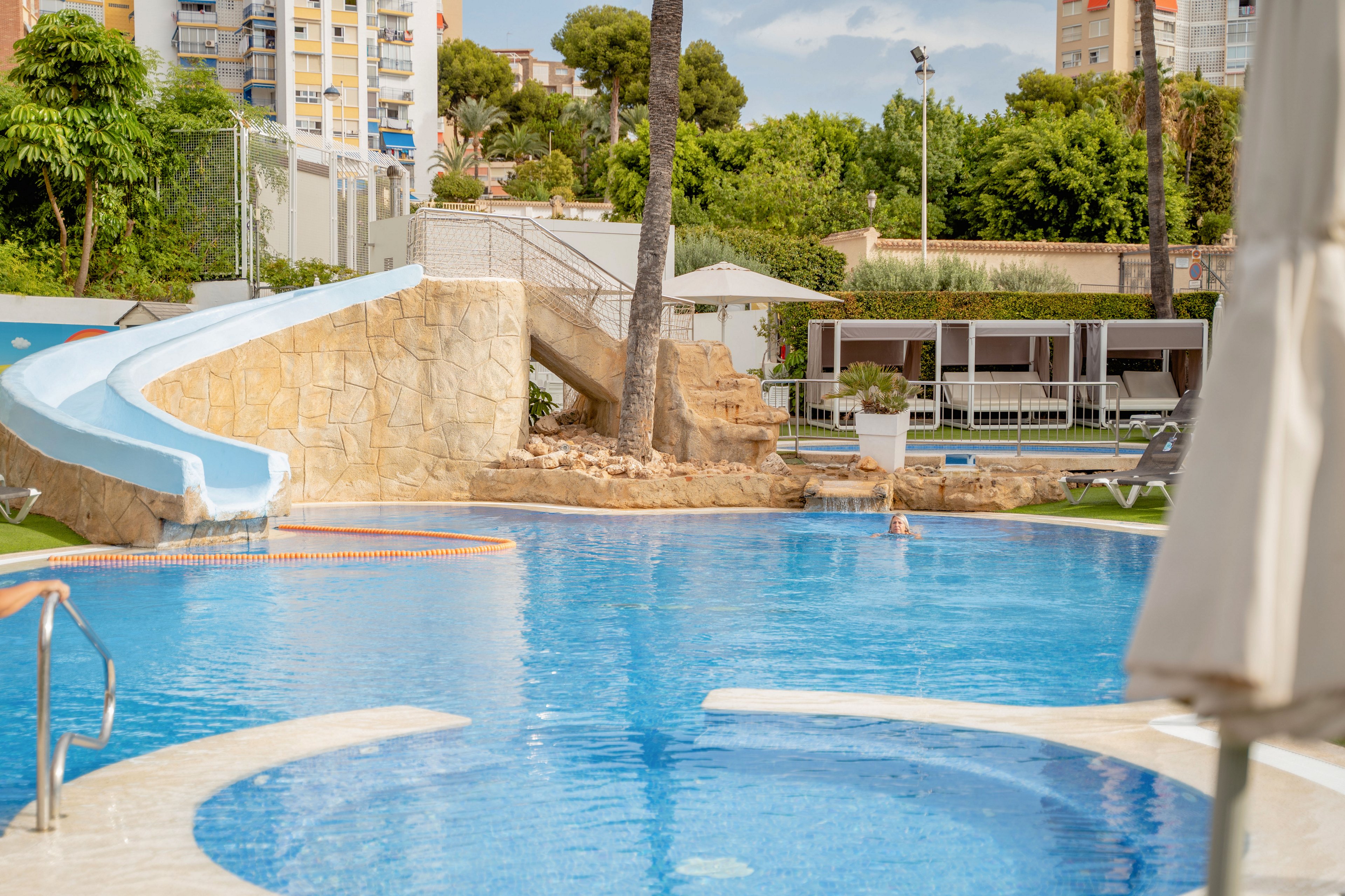 An outdoor resort swimming pool features a curved waterslide and stone steps leading down to the water. Lounge chairs and shaded cabanas line the poolside area, suggesting a relaxing vacation setting. Tall palm trees and lush greenery surround the pool, with apartment buildings visible in the background. The bright daylight and clear blue water create a cheerful, summery atmosphere.