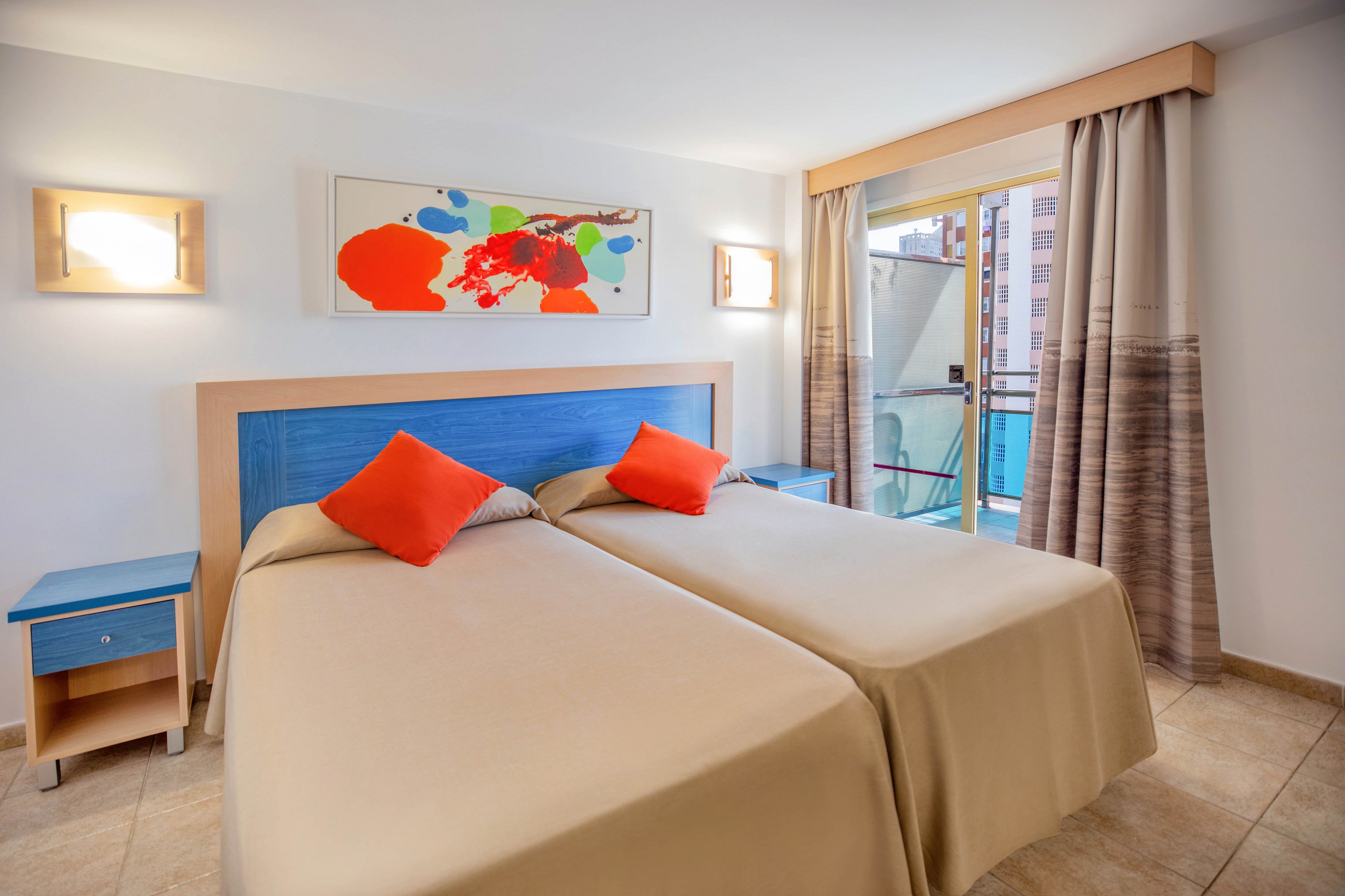 A clean, modern hotel room features two neatly made twin beds with beige bedspreads and bright orange accent pillows. The room includes a blue headboard, matching bedside tables, and warm wall-mounted lights. A colorful abstract painting hangs above the beds, adding a playful touch. Natural light enters through a glass door leading to a small balcony with city buildings visible outside.