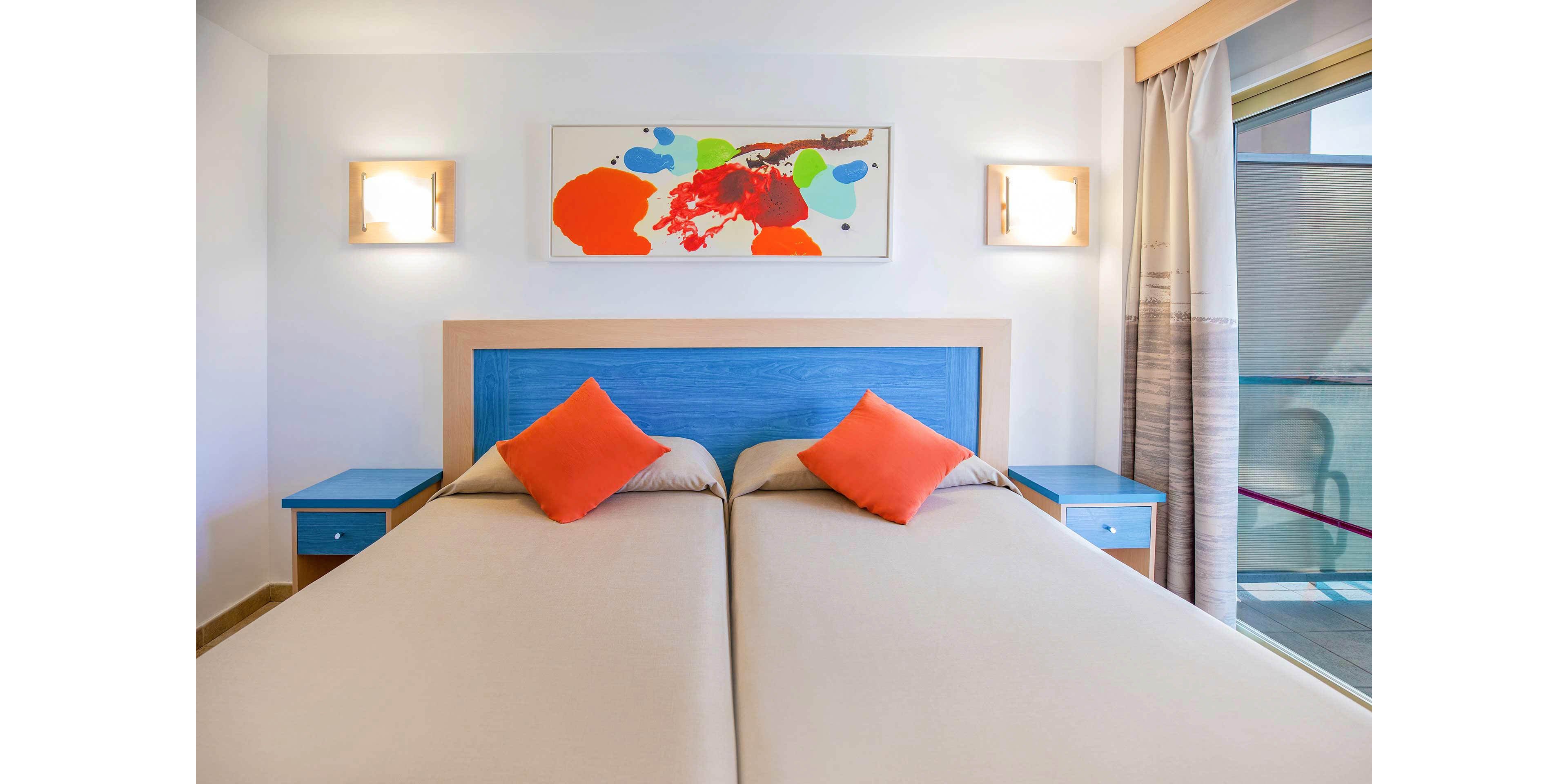 A modern hotel room features two neatly made twin beds pushed together, each with beige bedding and bright orange accent pillows. The headboard and bedside tables are painted in a vivid blue, adding contrast to the neutral walls. Abstract artwork with bold red, blue, and green splashes hangs above the bed, framed by two wall-mounted lights. A large window with sheer curtains allows natural light to brighten the clean, minimalist space.