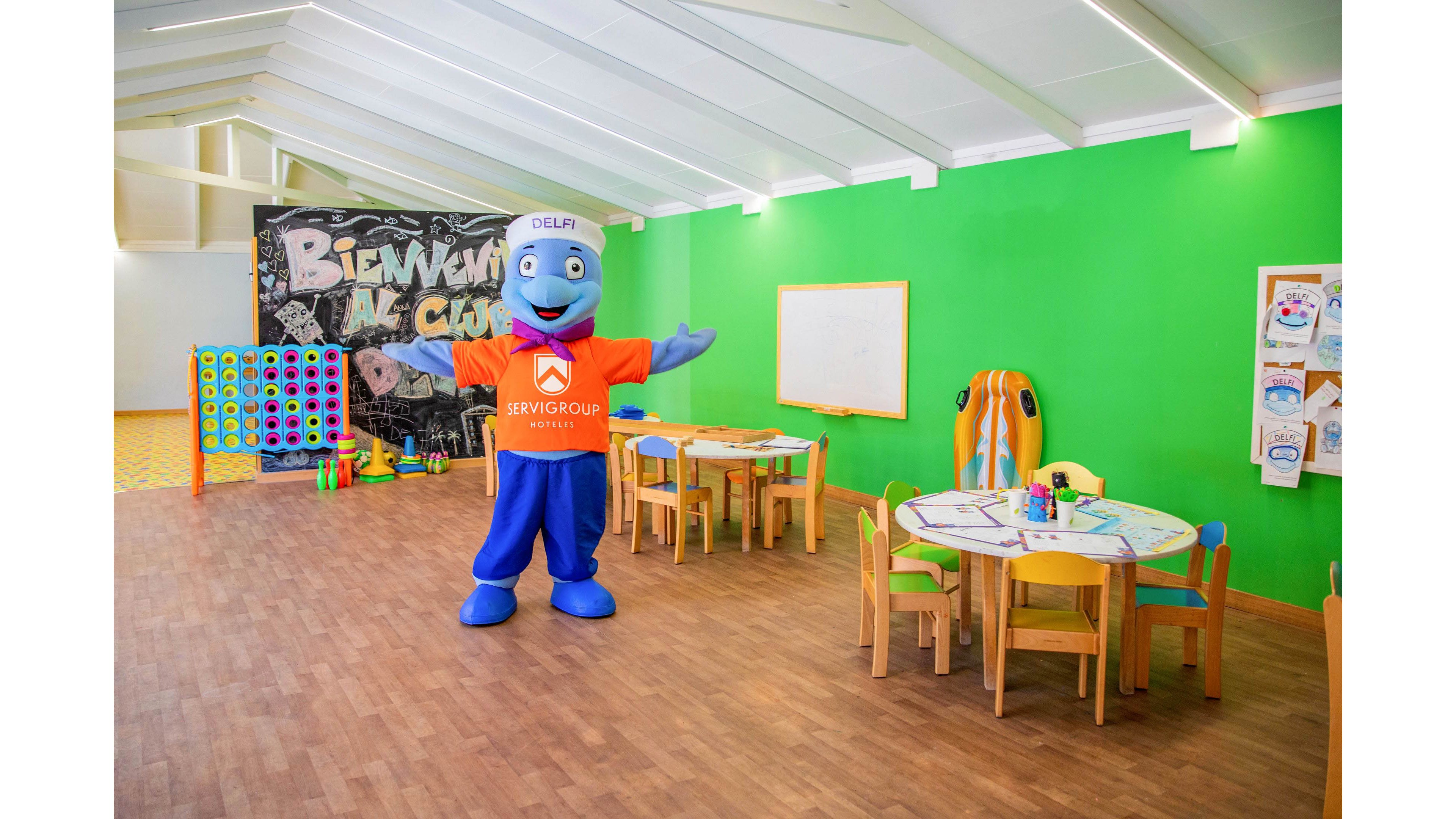 A cheerful blue dolphin mascot stands with open arms in a brightly lit children's playroom. The room features green walls, small wooden tables and chairs, and a large wall-mounted game board. A chalkboard-style mural in the background reads "BIENVENUE AU CLUB" in large letters. The playful setting suggests a welcoming environment for kids' activities and games.