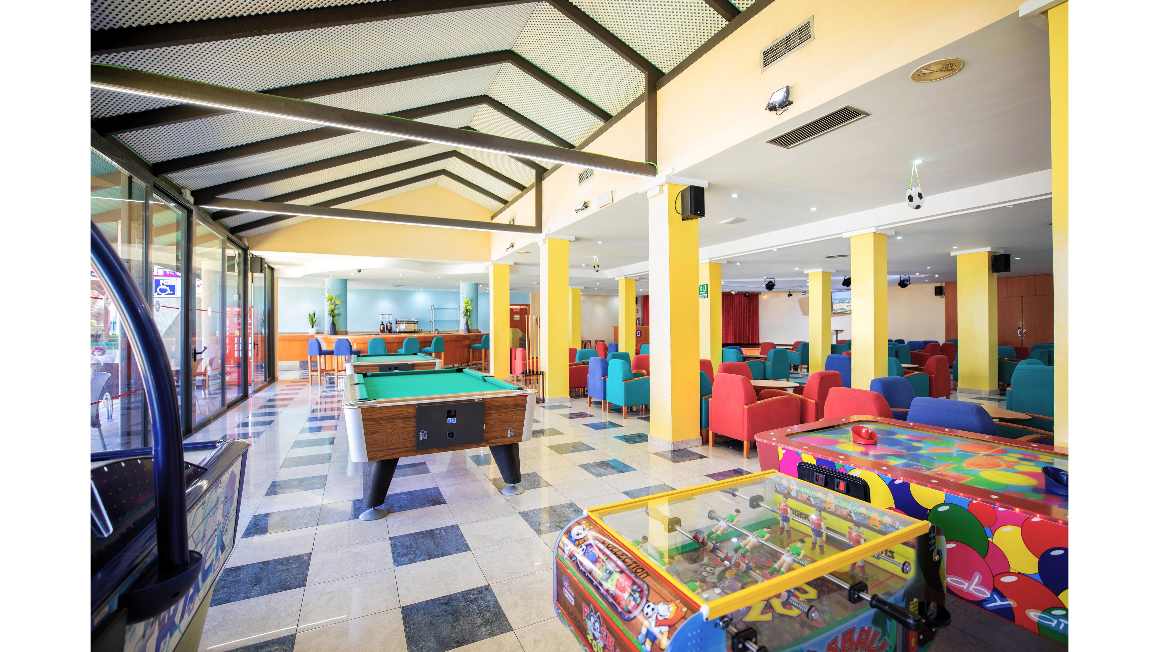 A spacious indoor game room features a central pool table surrounded by bright, colorful seating. Arcade machines and game tables line the sides of the room, adding to the playful atmosphere. Large windows and glass partitions let in natural light, while tiled floors and yellow columns create a clean, modern look. The space appears ready for guests to relax, socialize, and enjoy various games.
