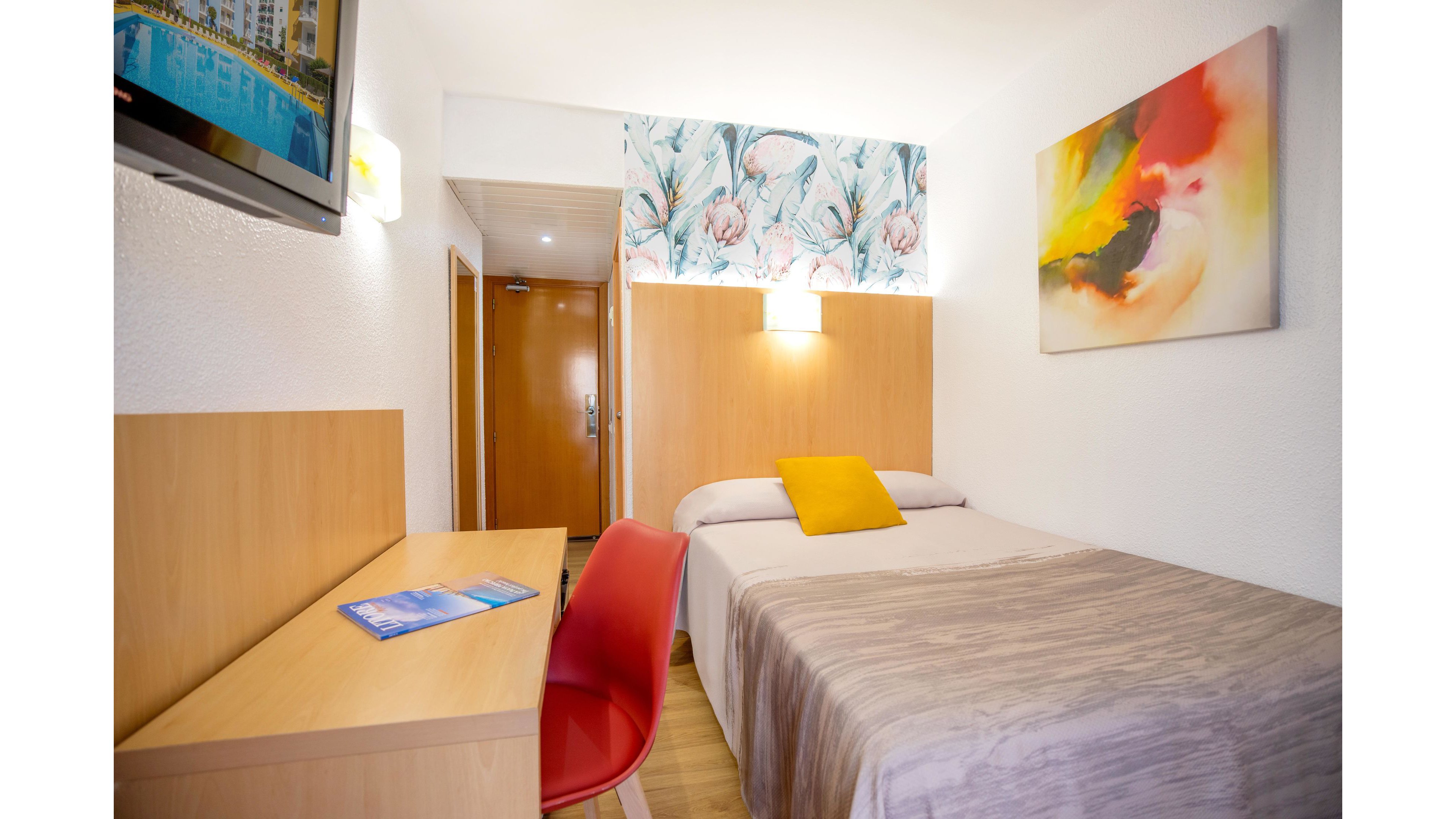 A small, neatly arranged hotel room features a single bed with a beige cover and a yellow accent pillow. A wooden desk with a red chair sits along one wall, holding a brochure or booklet. Colorful abstract artwork and floral wallpaper add visual interest above the bed. A wall-mounted television and warm lighting create a cozy, modern atmosphere in the compact space.