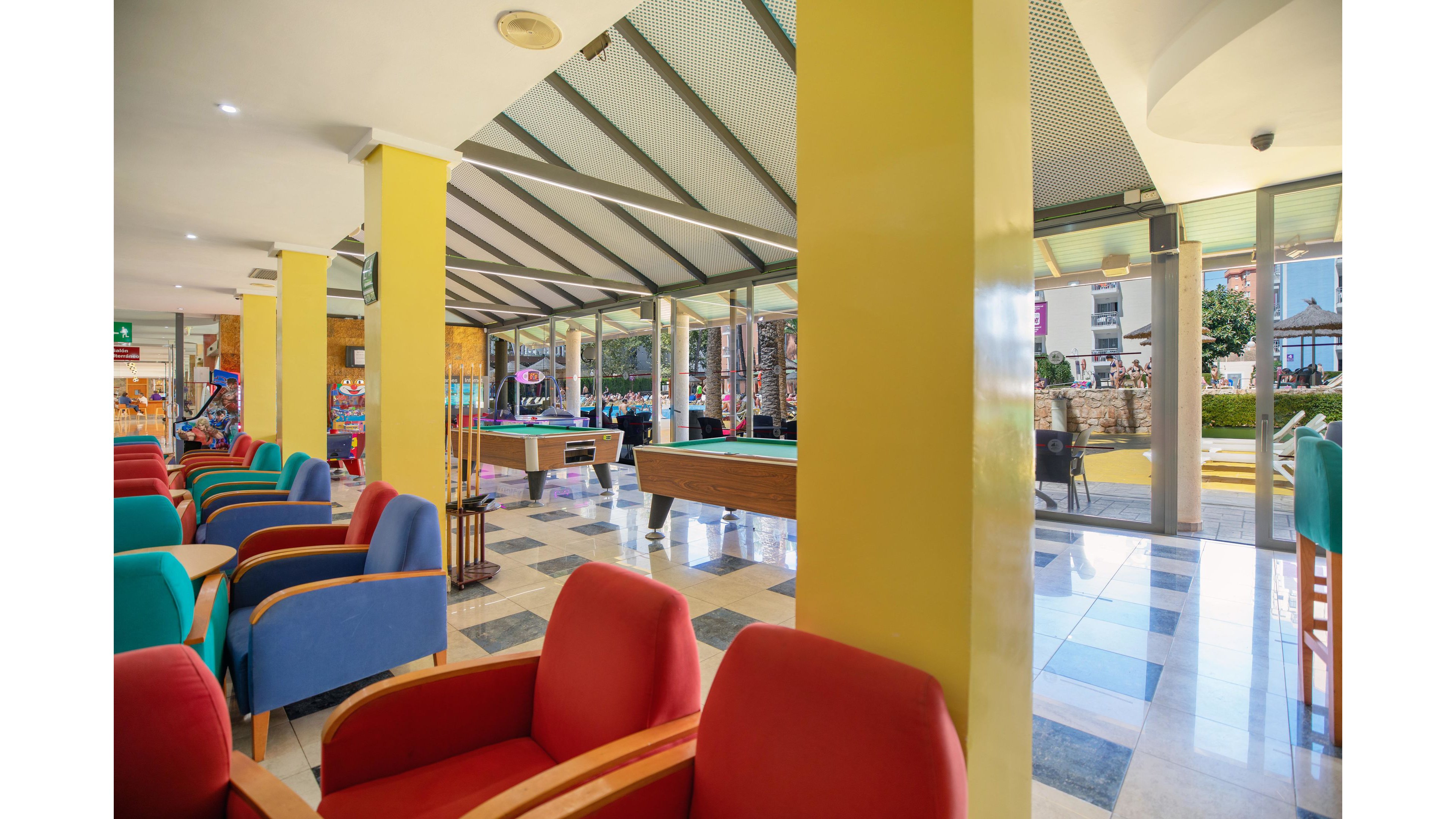 A bright hotel lounge features colorful armchairs arranged around low tables. In the center of the space, two pool tables stand on a checkered tile floor beneath a slanted glass roof. Large windows open to an outdoor area with palm trees and people walking by. The overall atmosphere is casual and inviting, suitable for relaxation and social games.