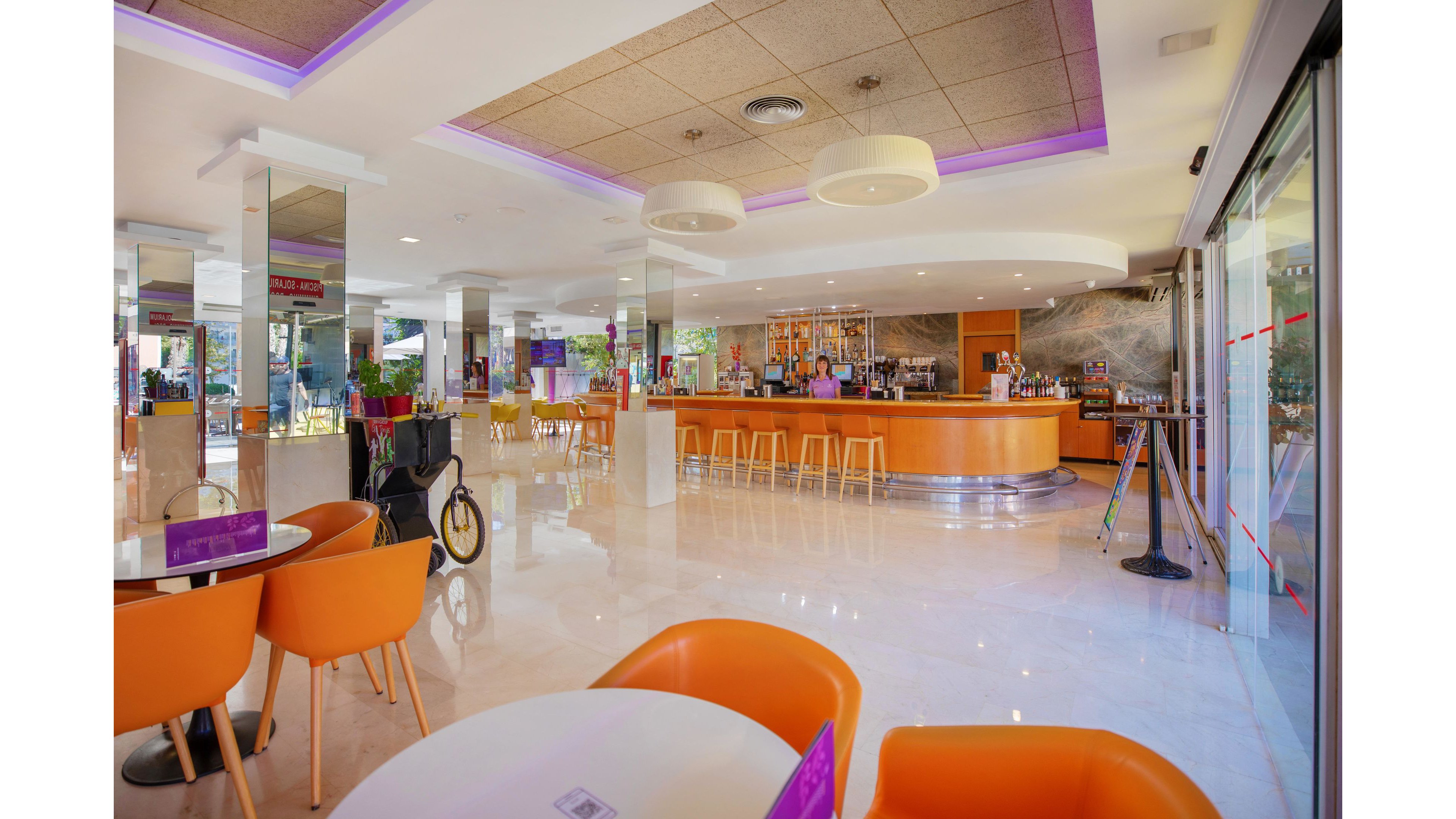 A spacious hotel bar interior features a curved wooden counter with high stools and a well-stocked back bar. Bright orange chairs and round white tables create a contemporary seating area on a glossy tiled floor. Large windows on the right let in natural light and offer views of greenery outside. Purple accent lighting in the ceiling and signage adds a colorful, modern atmosphere to the space.