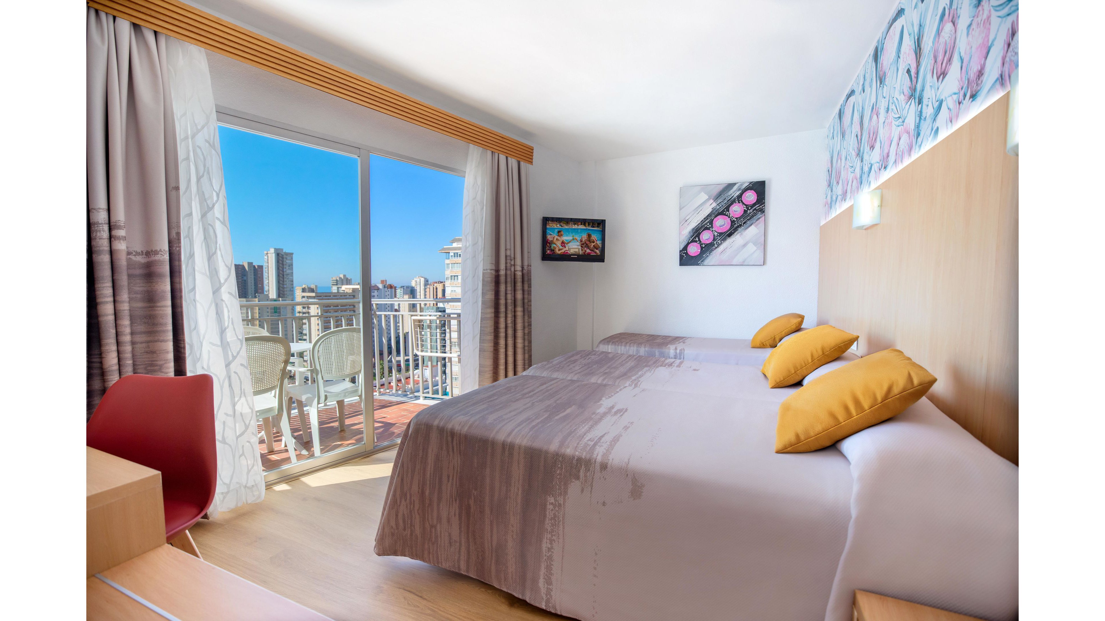 A modern hotel room features two neatly made beds with gray bedding and yellow accent pillows. Large sliding glass doors open onto a balcony with white chairs, overlooking a sunny cityscape of tall buildings and blue sky. Light wood furniture, a red chair, and patterned curtains add contemporary touches to the interior. Wall art and a small mounted television complete the cozy, inviting atmosphere.