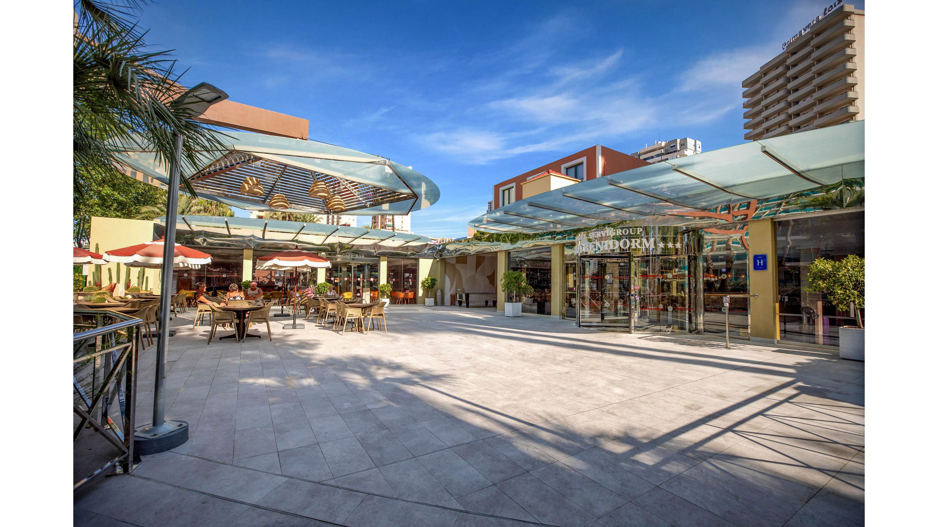 A contemporary open-air shopping plaza features glass-fronted stores and outdoor seating areas. Tables with umbrellas line the left side, suggesting a café or restaurant setting under clear blue skies. Modern glass canopies and clean tiled flooring create a bright, upscale atmosphere. A few people are visible in the distance, adding a sense of casual activity to the scene.