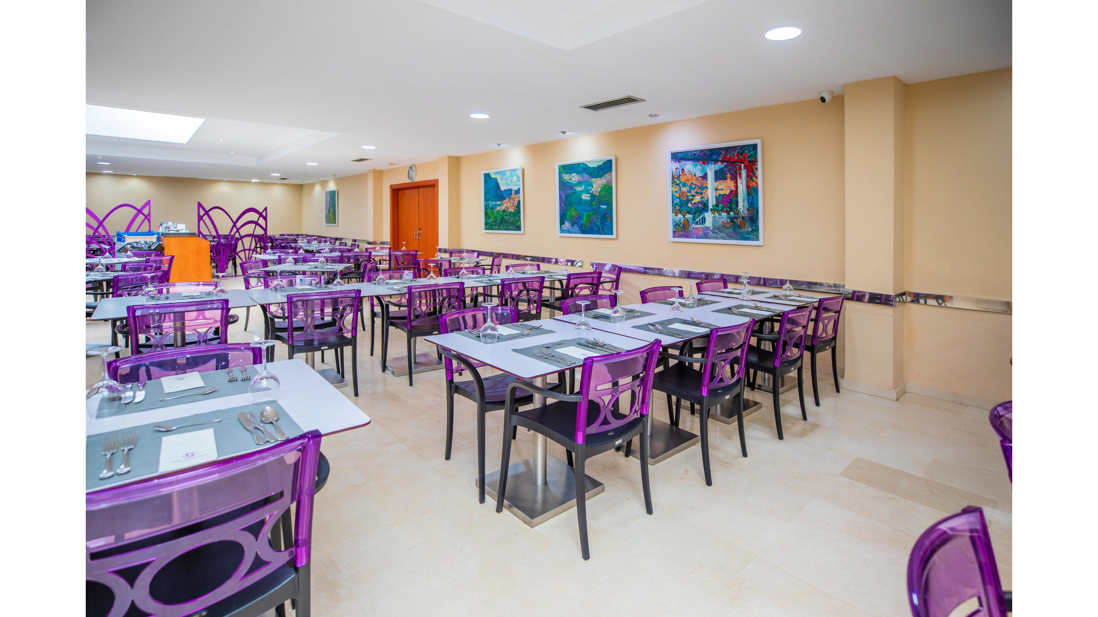 A spacious cafeteria is arranged with rows of tables and transparent purple chairs. Each table is neatly set with placemats, cutlery, glasses, and napkins, ready for diners. Colorful paintings hang on the beige walls, adding visual interest to the clean, contemporary interior. The room is brightly lit with ceiling lights and natural light, creating an inviting dining atmosphere.