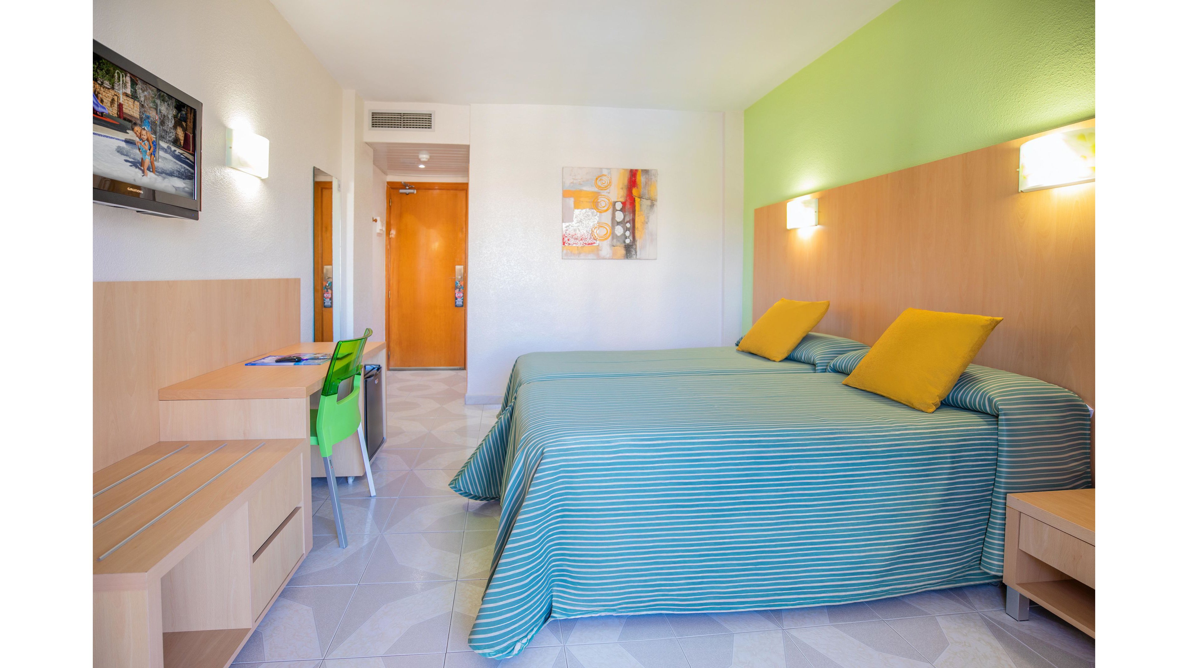 A colorful hotel room features two neatly made twin beds with striped blue bedspreads and yellow accent pillows. The space includes a light wood desk with a green chair, a wall-mounted television, and a small bench. Soft wall lights and a green accent wall create a cheerful, contemporary atmosphere. A wooden door and abstract artwork complete the clean, modern interior design.
