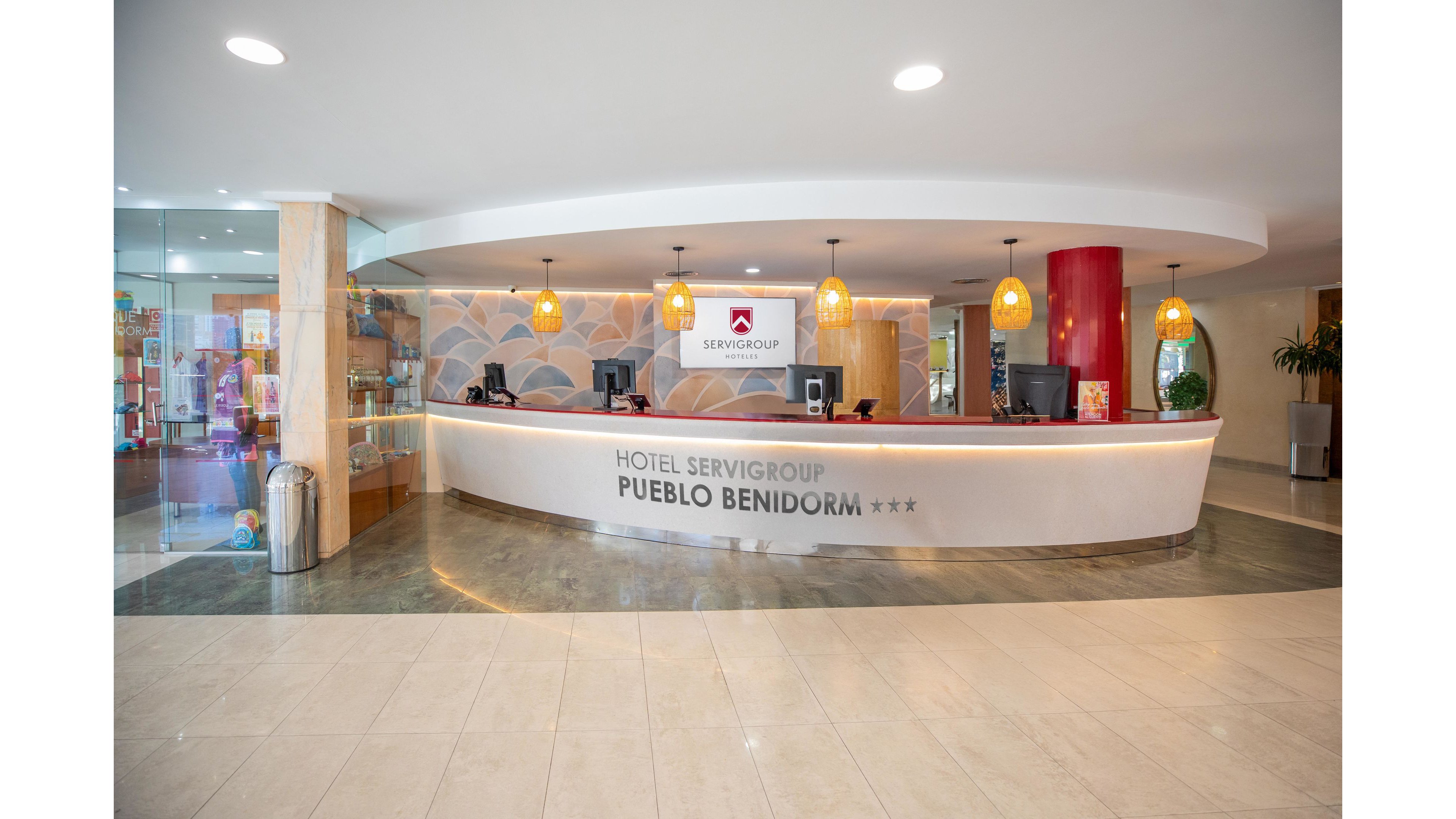 Spacious hotel reception area featuring a curved white front desk with multiple computer stations. The setting appears bright and welcoming, with pendant lights, glass display shelves, and polished tile flooring. Text on the desk reads "HOTEL SERVIGROUP PUEBLO BENIDORM" followed by three stars. The background includes decorative wall art and a red column, enhancing the contemporary interior design.