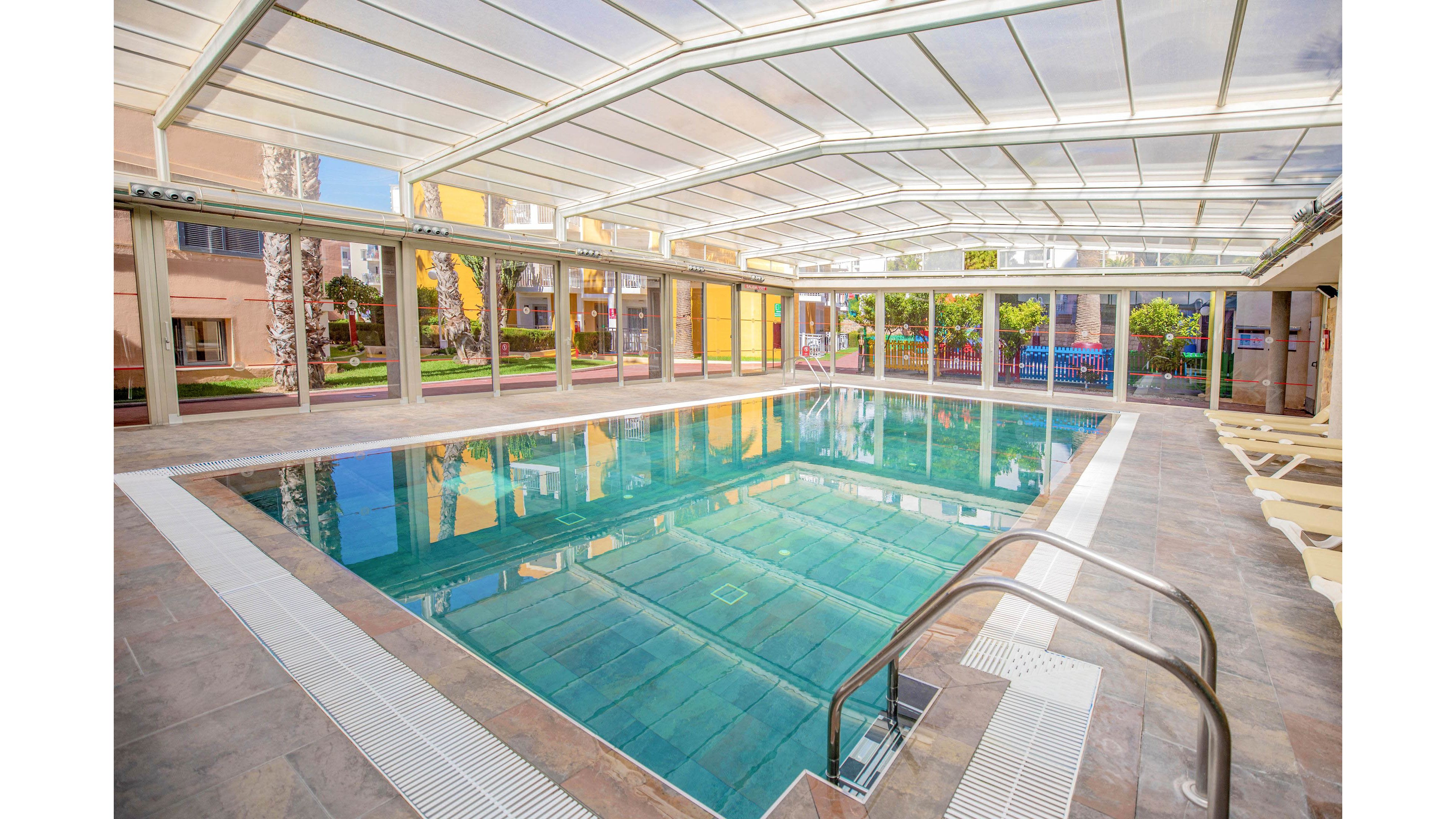 A clear turquoise swimming pool sits inside a spacious glass-enclosed structure. Sunlight streams through the transparent roof and walls, reflecting off the calm water surface. Lounge chairs line the tiled deck, suggesting a relaxing recreational environment. Buildings and greenery are visible outside the enclosure, adding a resort-like atmosphere.