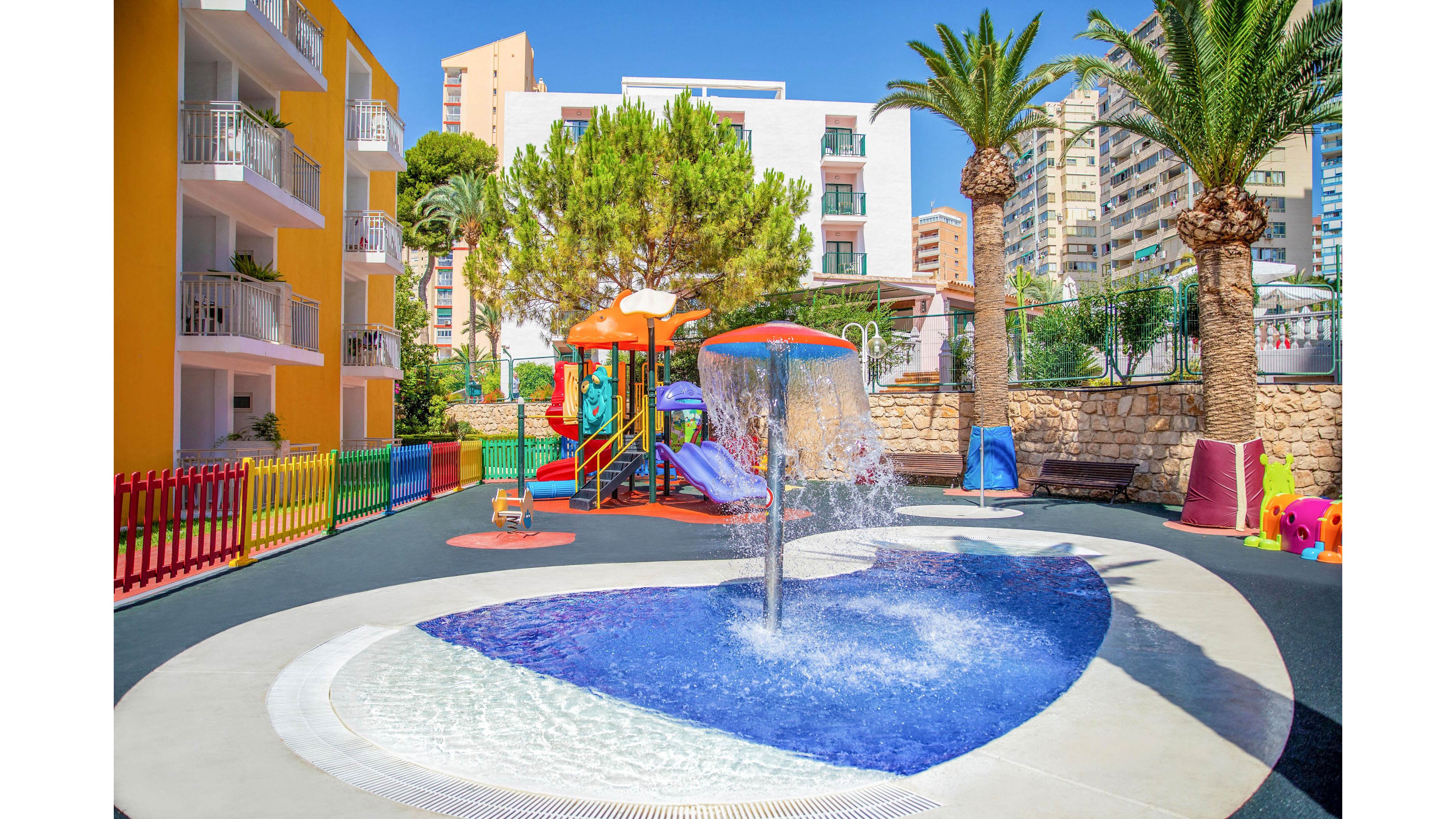 An outdoor splash pad with a central water feature and shallow pool is set in a brightly colored hotel courtyard. A small playground with slides and climbing structures stands nearby, surrounded by palm trees and modern apartment buildings. The ground is covered with soft safety surfacing and bordered by a multicolored fence. The scene is sunny and vibrant, suggesting a family-friendly vacation destination.