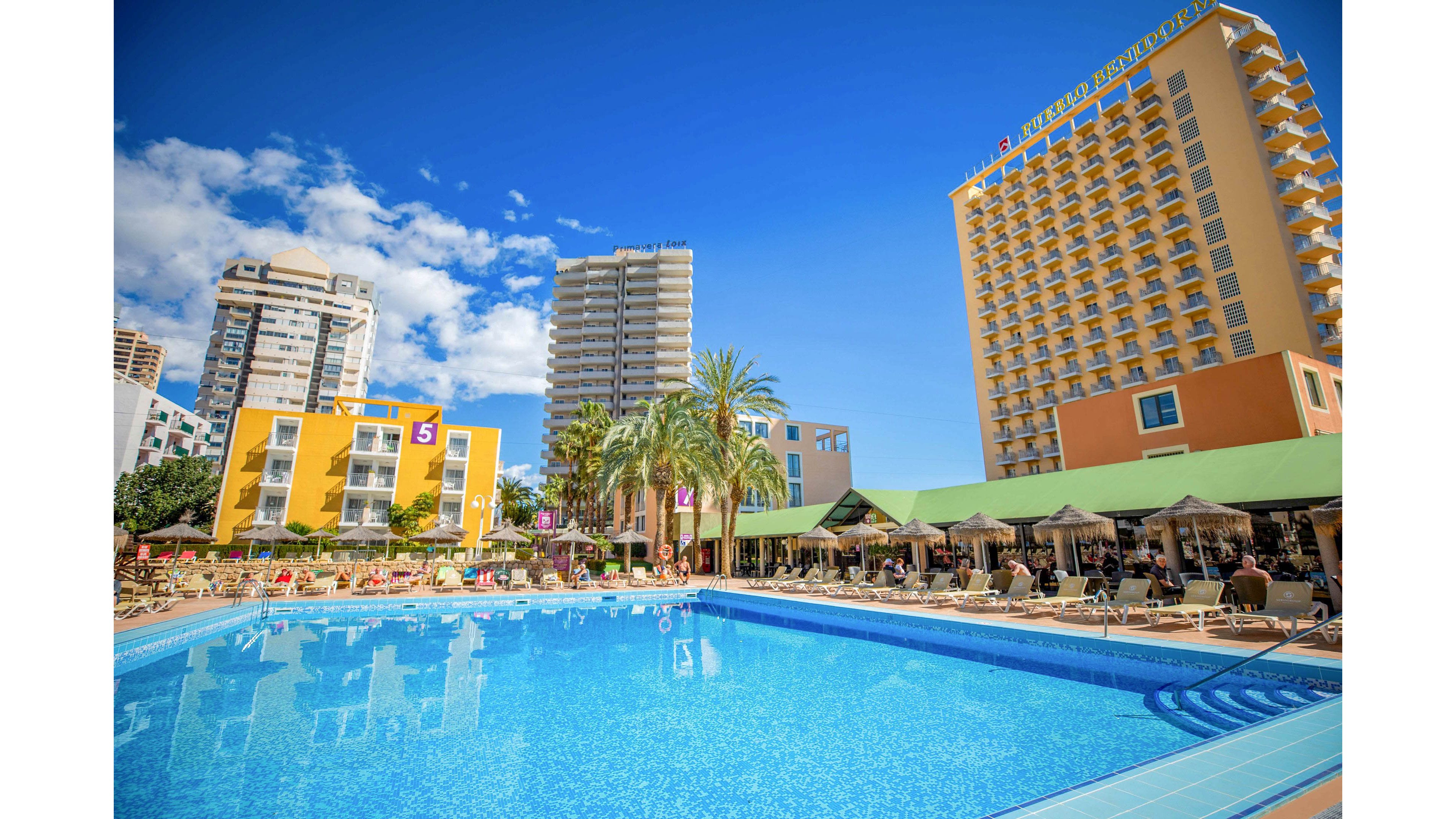 A large outdoor swimming pool reflects the bright blue sky at a sunny resort hotel. Guests relax on sun loungers under green canopies around the pool area. Tall modern hotel buildings and palm trees frame the scene, creating an urban vacation atmosphere. The overall mood is vibrant, warm, and inviting, suggesting a lively holiday destination.