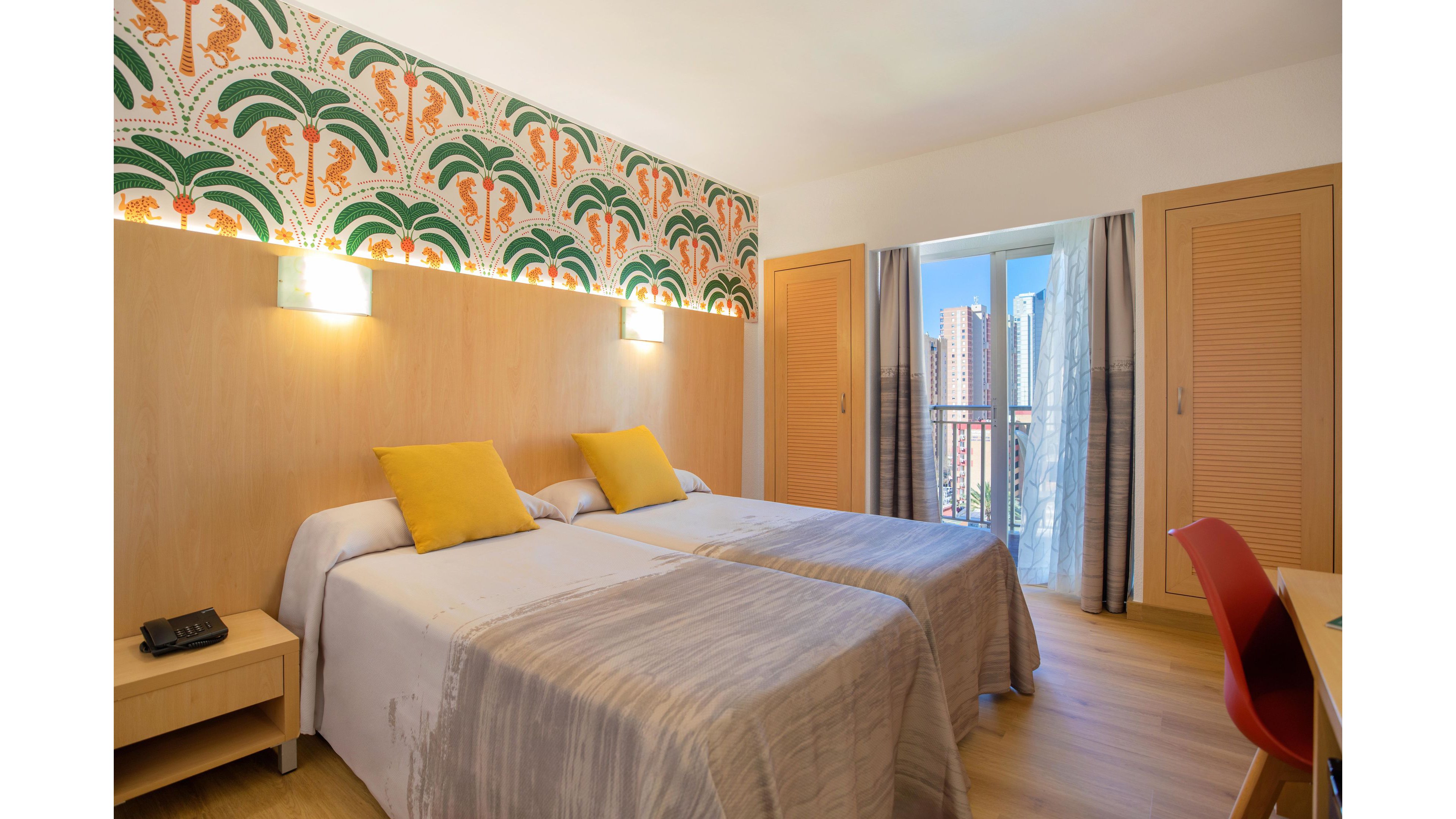 A modern hotel room features two neatly made twin beds with light gray bedspreads and yellow accent pillows. The space is decorated with a colorful palm tree patterned wall panel above a light wood headboard. Natural light enters through a glass door leading to a small balcony overlooking tall city buildings. Built-in wooden wardrobes, a bedside table with a phone, and a red chair complete the cozy, contemporary interior.