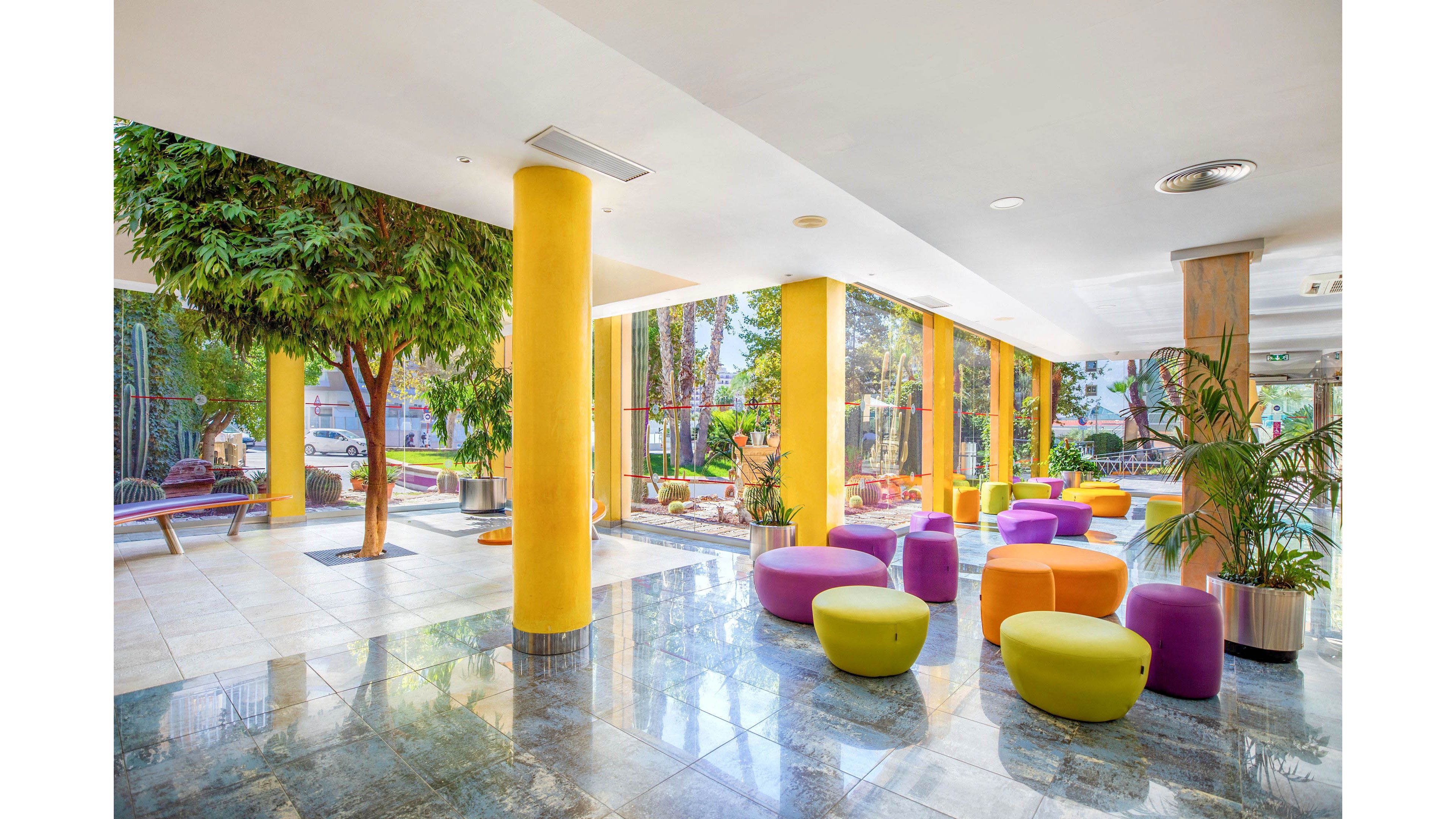 Bright, contemporary lobby interior featuring bold yellow columns and colorful round ottomans. Large floor-to-ceiling windows open onto a lush outdoor garden with palm trees and greenery. The polished tile floor reflects natural light, enhancing the airy, spacious feel of the room. Potted plants and indoor trees add a relaxed, resort-like atmosphere to the modern space.