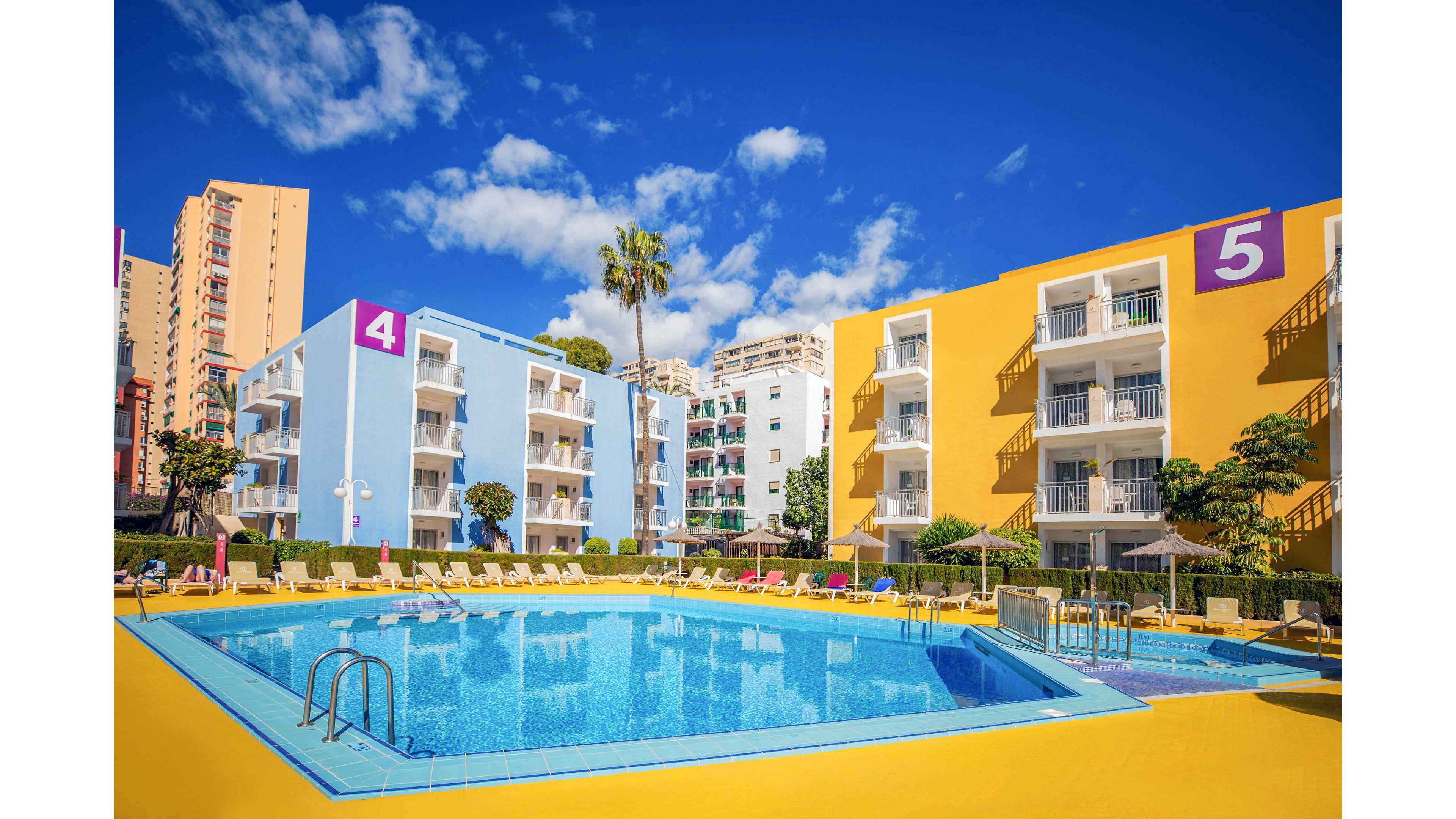A bright resort complex features a large outdoor swimming pool surrounded by yellow decking and sun loungers. Low-rise apartment buildings painted in blue and yellow frame the pool area under a vivid blue sky. The buildings are marked with large purple and white numbers 4 and 5 on their corners. Tall palm trees and distant high-rise structures add to the sunny holiday atmosphere.