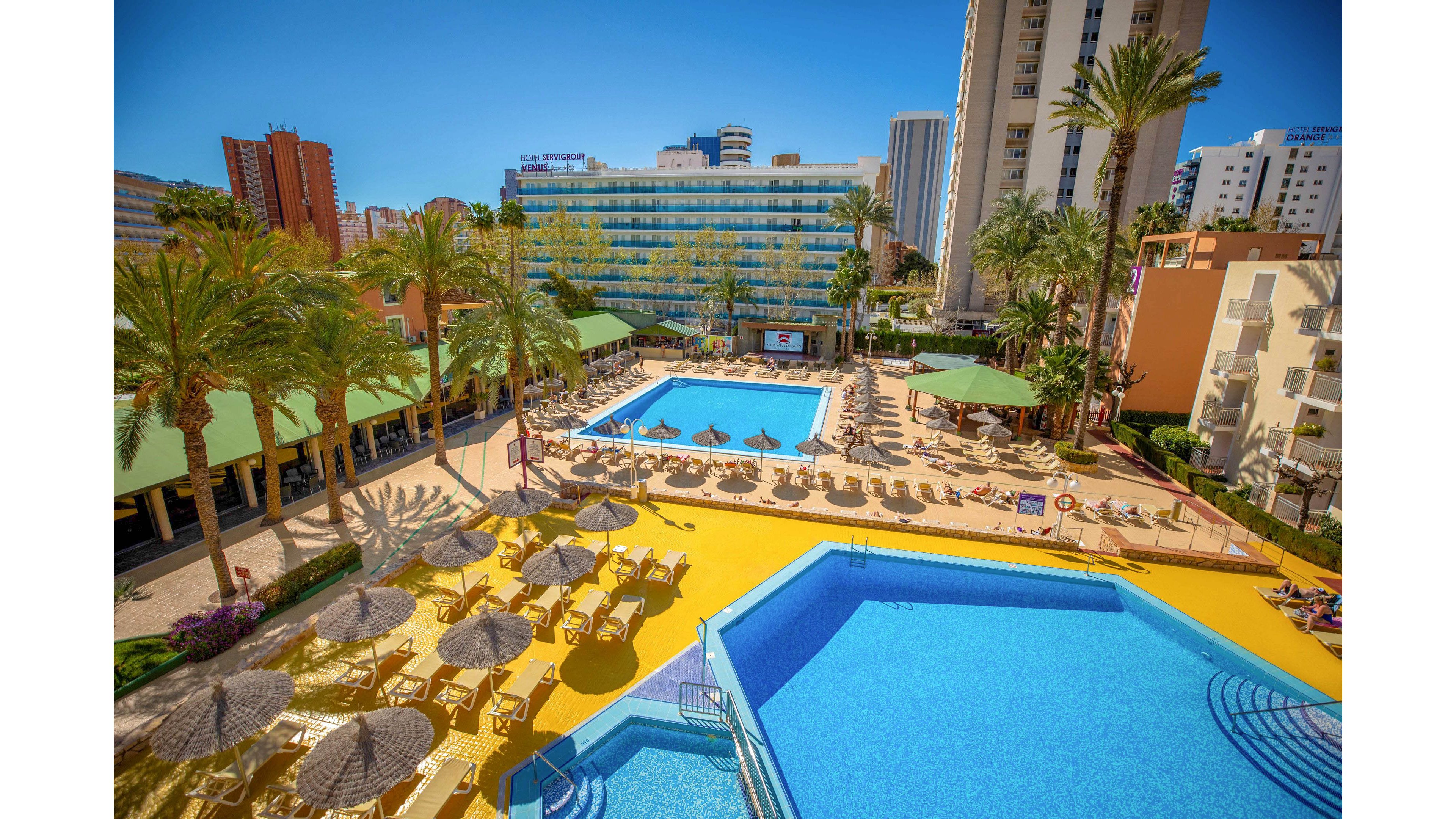 An elevated view shows a vibrant resort hotel complex with two large outdoor swimming pools. Sun loungers and straw parasols surround the pools on bright yellow terraces, creating a lively vacation atmosphere. Tall palm trees and green-roofed structures frame the scene, while high-rise buildings and a clear blue sky rise in the background. Guests relax around the water, suggesting a warm, sunny holiday destination in an urban coastal area.