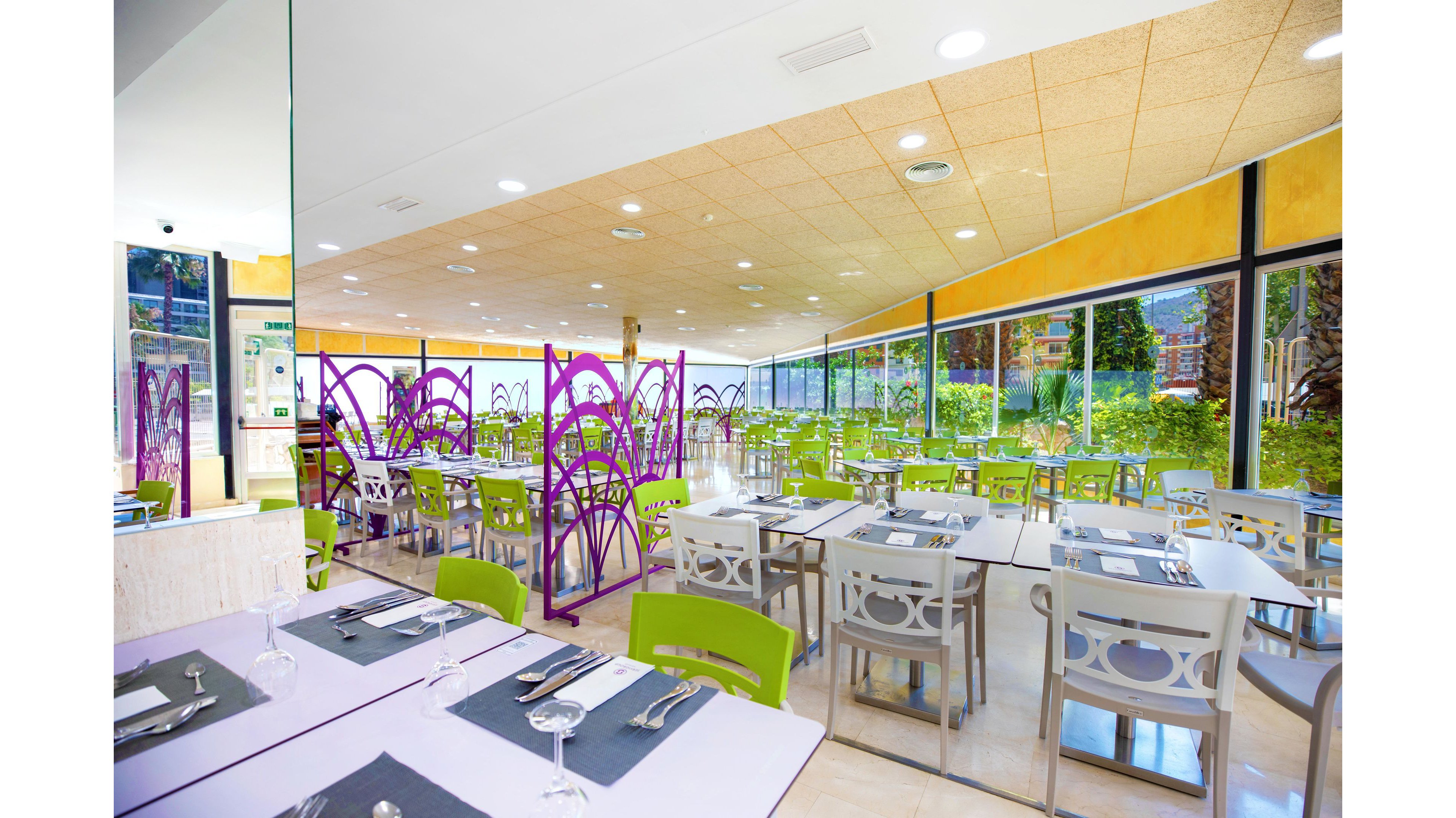 A spacious, modern cafeteria is filled with neatly arranged tables and chairs. Lime green and gray chairs surround square tables set with placemats, cutlery, and glassware. Large floor-to-ceiling windows line one side, offering views of greenery and letting in abundant natural light. Purple decorative partitions add a vibrant accent to the clean, contemporary interior.