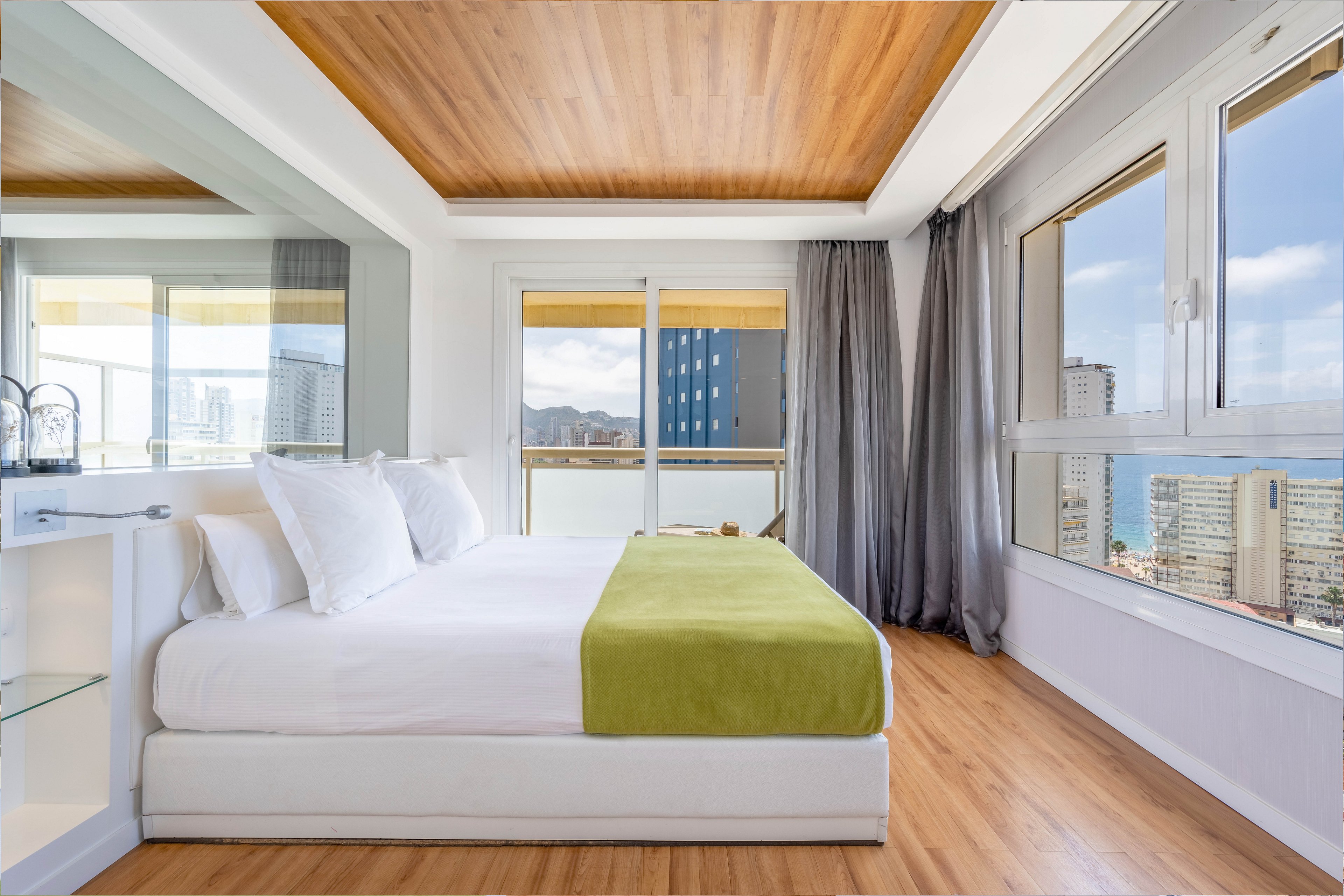 A modern, minimalist bedroom features a white platform bed with crisp white linens and a green throw blanket. Large windows and a glass door fill the room with natural light and reveal a high-rise cityscape and distant sea. The space includes light wood flooring, a wood-paneled ceiling, and sheer gray curtains framing the view. Clean lines and neutral tones create a calm, contemporary atmosphere ideal for relaxation.