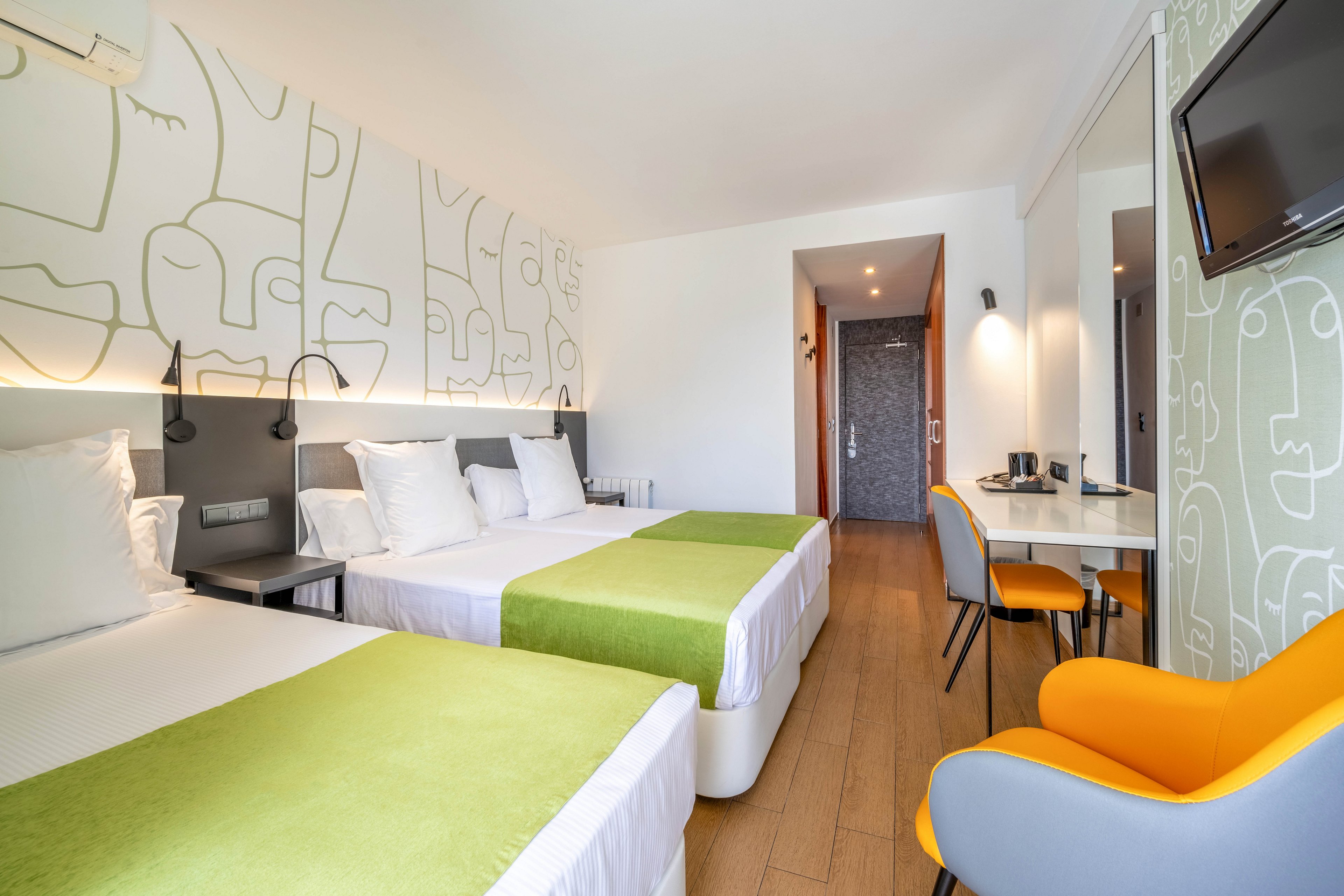 Contemporary hotel room featuring two neatly made twin beds with white linens and green runners. The space includes a work desk with chair, wall-mounted television, and a bright yellow armchair. Minimalist wall art with abstract faces decorates the headboard wall and side wall. Warm wood flooring and recessed lighting create a clean, inviting atmosphere suitable for travelers or business guests.