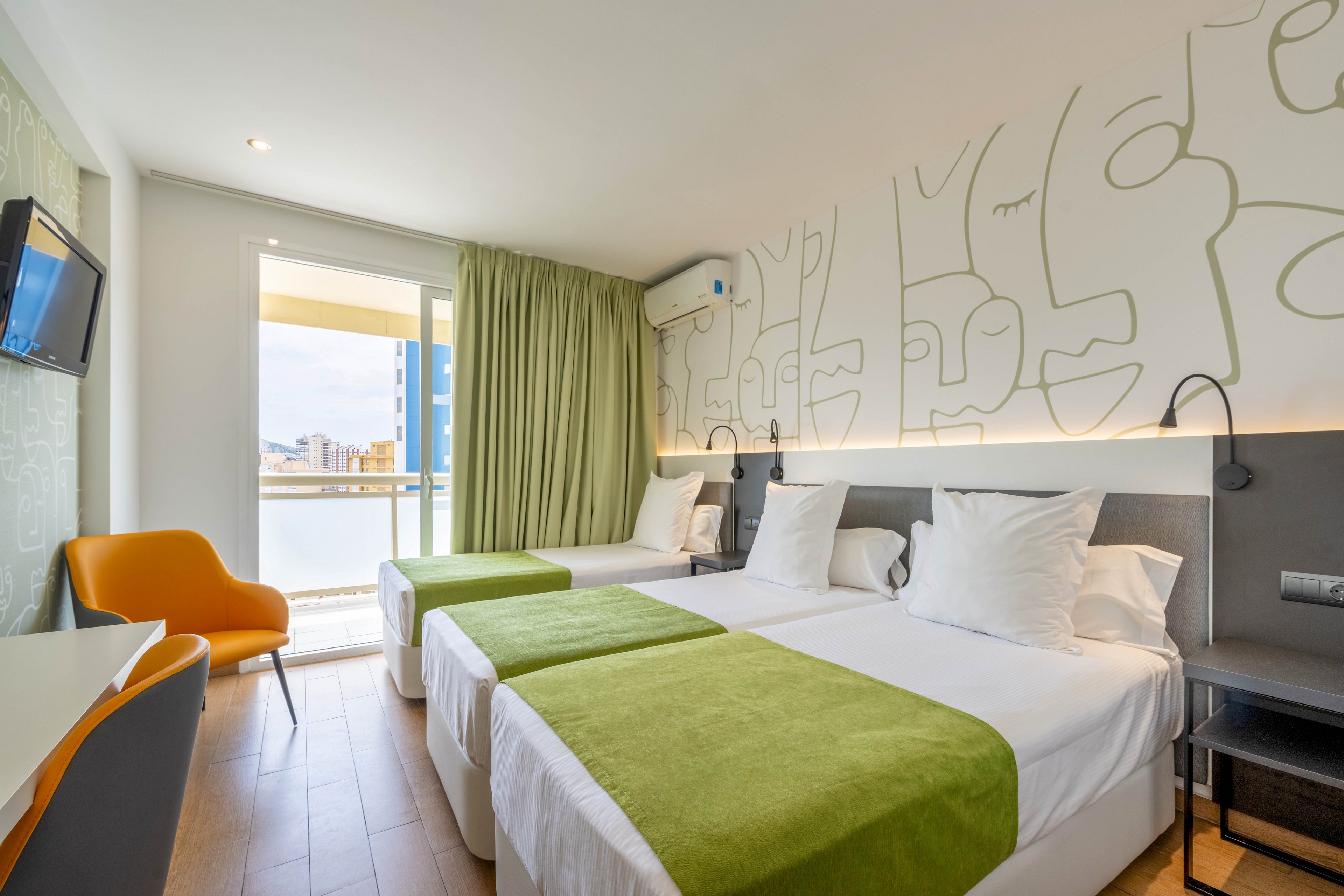 Bright contemporary hotel room featuring two neatly made twin beds with green runners and white linens. The space includes a wall-mounted television, a white desk, and an orange accent chair. Large sliding glass doors open to a balcony with a city and waterfront view, allowing natural light to fill the room. Minimalist wall art and soft green curtains contribute to the clean, modern aesthetic.
