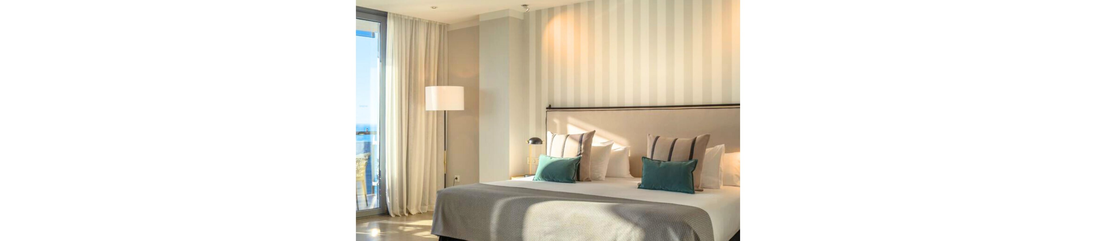 A contemporary hotel bedroom features a large neatly made bed with decorative pillows and a padded headboard. Soft natural light streams in from a glass balcony door, illuminating the neutral striped wall and light-colored curtains. A floor lamp and bedside table with a small reading lamp add to the cozy, inviting atmosphere. The overall design is minimal, bright, and elegant, suggesting a relaxing vacation stay.