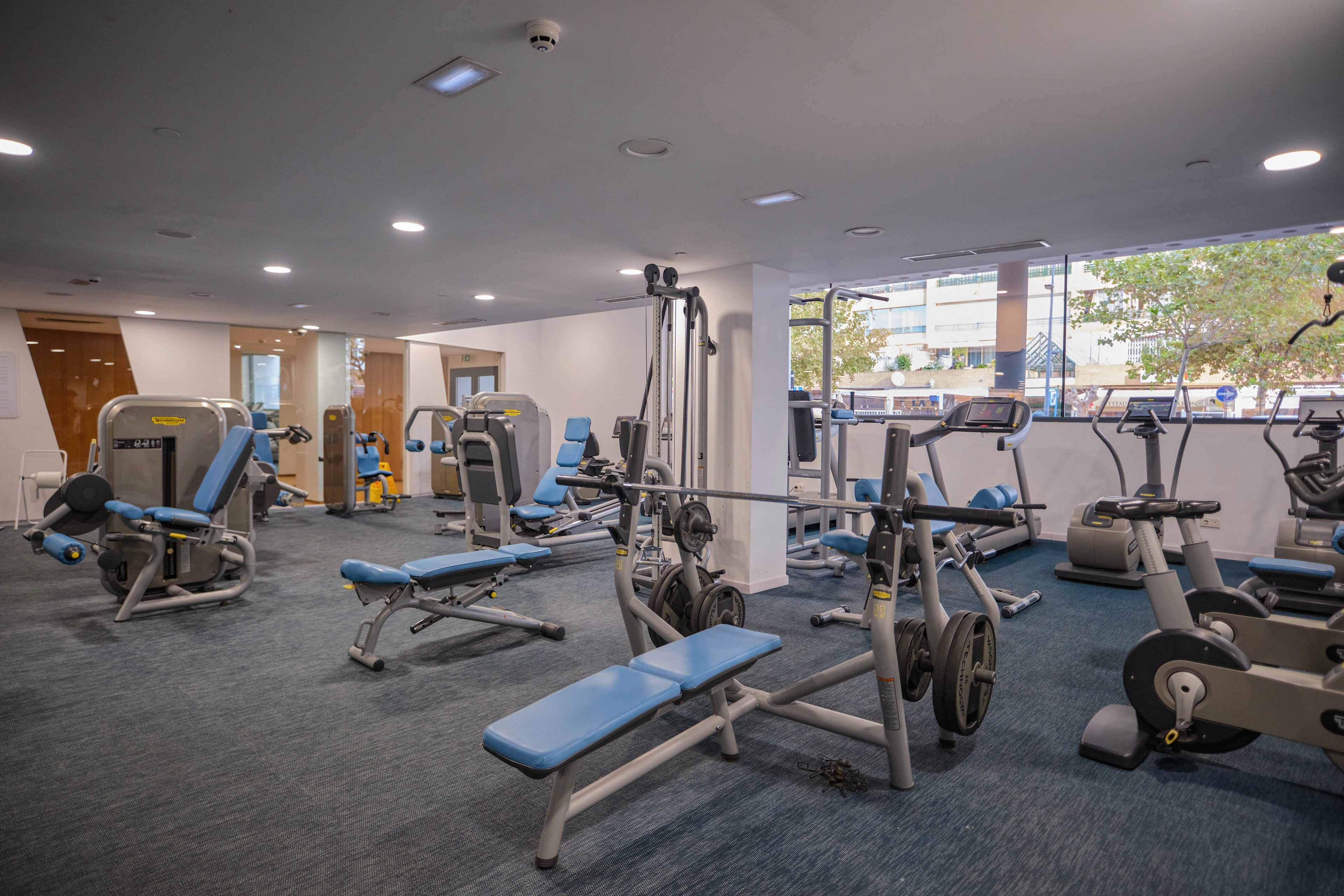 A modern fitness room features various exercise machines and weight benches neatly arranged on a blue carpeted floor. The space includes cardio equipment, strength-training machines, and adjustable benches with blue padding. Large windows along one wall let in natural light and reveal an urban street scene outside. The gym appears clean, organized, and ready for individual workouts.