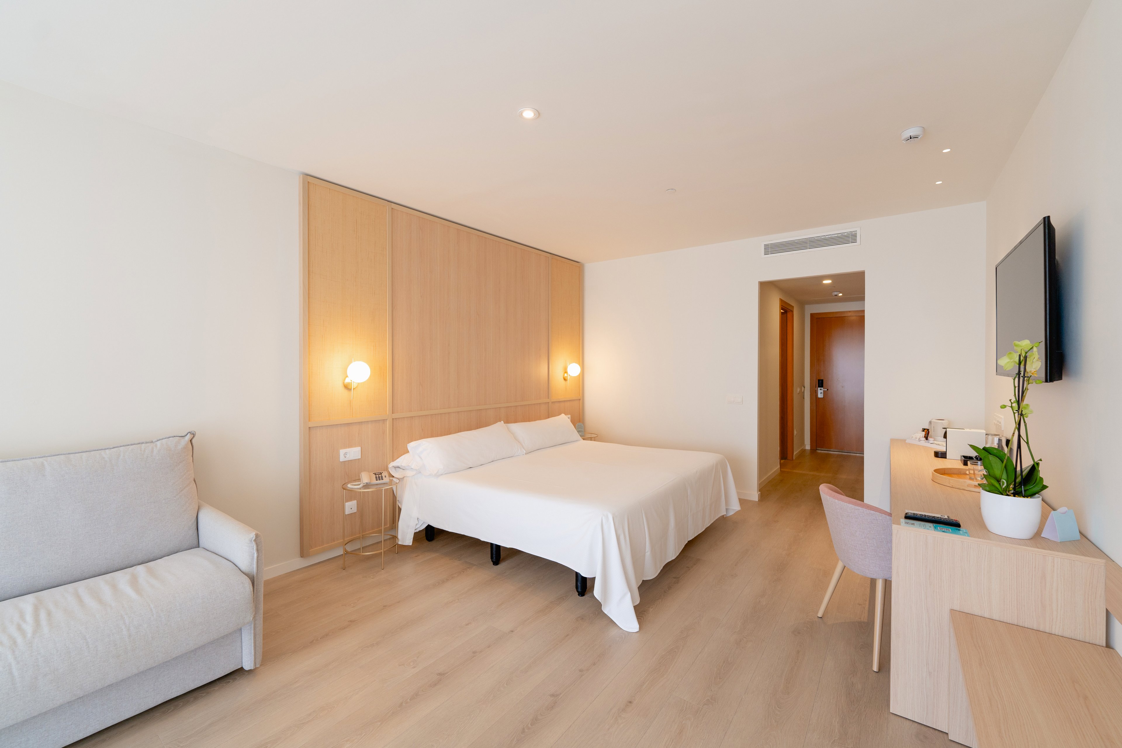 A spacious and well-lit hotel room featuring a double bed with white linens, a sofa, and a wooden desk. The room has a minimalist design with light wood accents and neutral tones. A potted plant and wall-mounted TV add to the contemporary aesthetic. The setting is clean and inviting, ideal for relaxation or work.