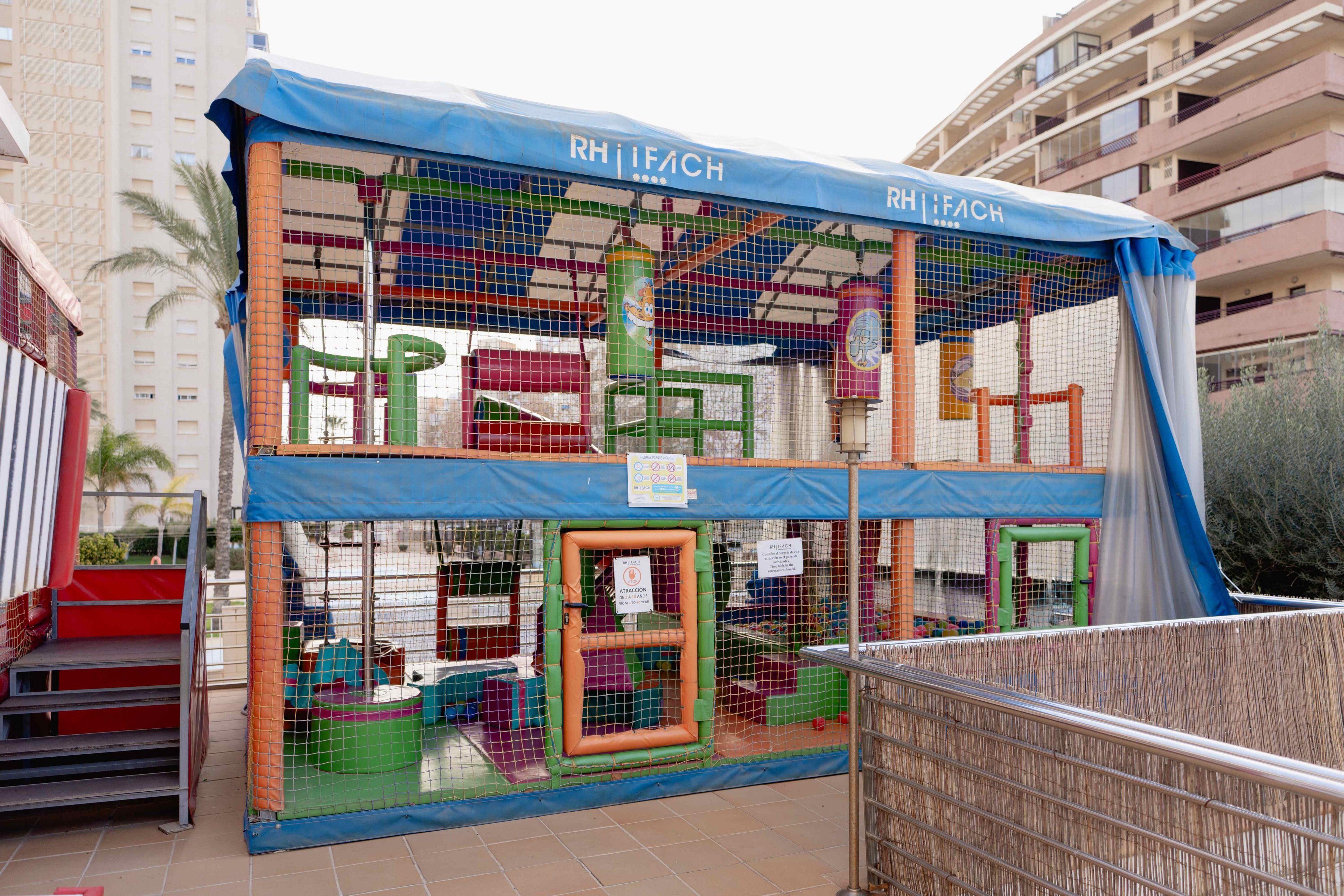 A colorful two-level soft play structure stands outdoors, enclosed with safety netting and covered by a blue canopy labeled RH IFACH. The play area features padded frames, tunnels, and climbing elements in bright green, red, yellow, and purple. It is situated on a tiled terrace between residential or hotel buildings, suggesting a recreational space for families. Signs are posted on the netting, indicating rules or information for visitors.