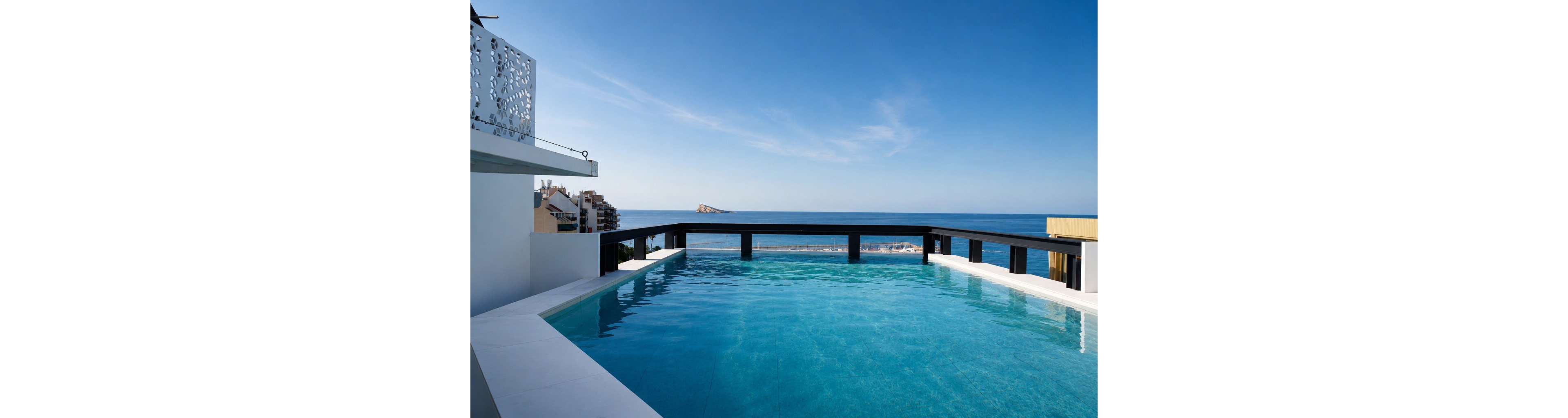 A modern infinity rooftop pool stretches toward a clear blue horizon, seamlessly blending with the distant sea. Contemporary buildings and apartments line the coastline below, adding an urban touch to the coastal setting. The calm water, minimalist architecture, and bright daylight create a serene, luxurious atmosphere. The scene emphasizes relaxation, vacation living, and panoramic ocean views.