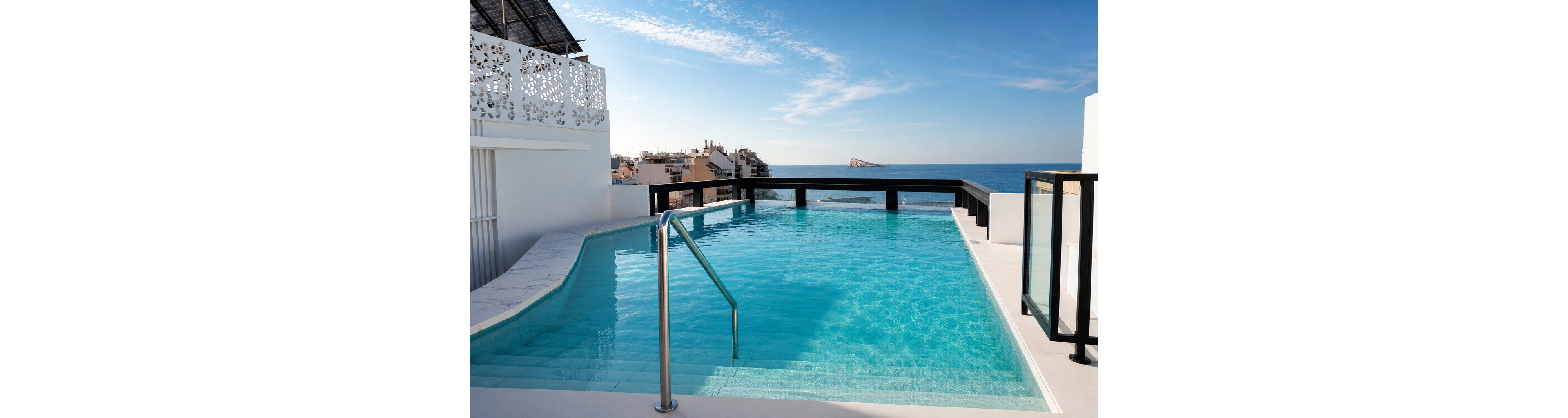 A modern rooftop infinity pool stretches toward a distant coastal skyline and open sea. Clear turquoise water fills the pool, with steps leading down and a metal handrail providing access. Minimalist white architecture and dark railings frame the view, while scattered buildings and a rocky islet appear on the horizon. The bright daylight and blue sky create a serene, luxurious vacation atmosphere.