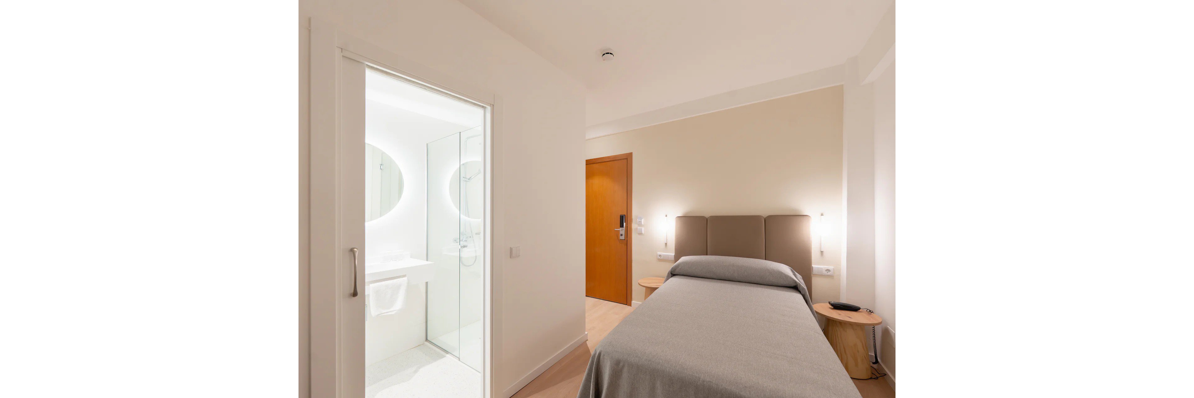 A compact hotel room features a single bed with a neutral gray bedspread and a padded headboard. The space is bright and minimalist, with white walls and light wood flooring. An open door reveals an ensuite bathroom with a round illuminated mirror and a white towel hanging neatly. Soft wall-mounted lights and a wooden nightstand contribute to the clean, modern aesthetic.