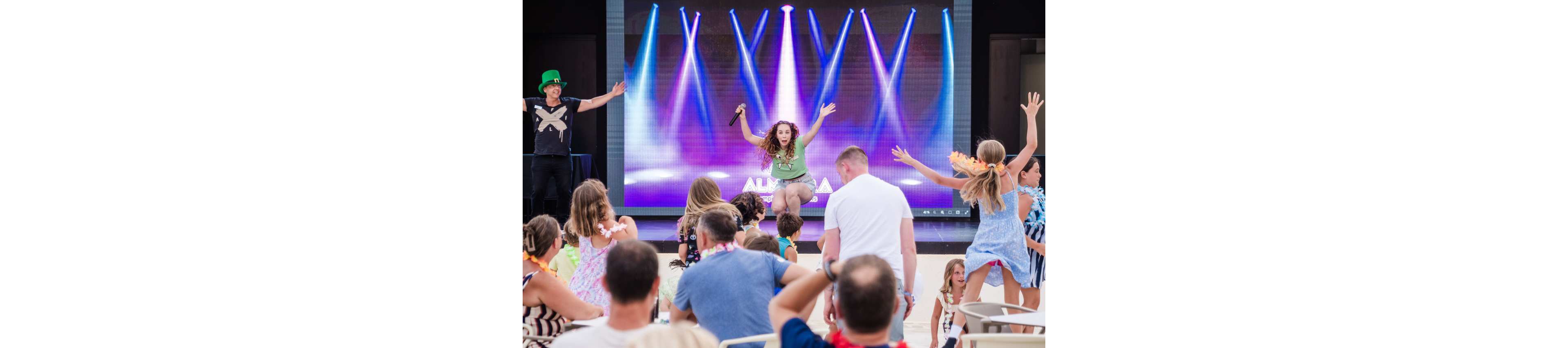 A female performer with a microphone leads a group of children and adults in a dance in front of a large digital stage screen. Colorful spotlights and beams of light are displayed on the screen, creating a concert-like atmosphere. The audience, mostly families with young kids, raises their arms and follows the entertainer’s movements. The event appears to take place outdoors in daylight, giving the scene a festive holiday vibe.