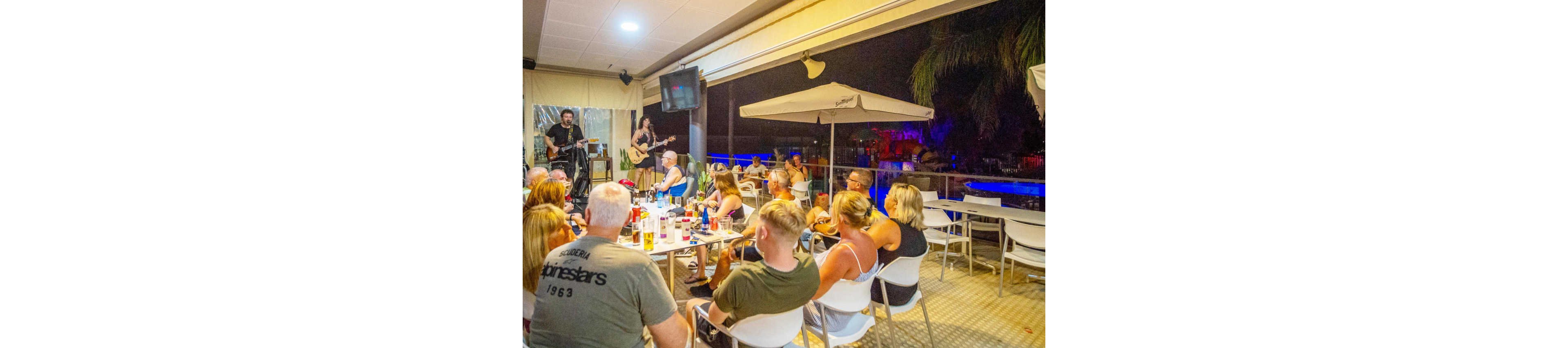 A group of adults sit at tables in an outdoor bar or restaurant at night, watching a live band perform. The scene is brightly lit under a covered terrace with a view of a pool or illuminated area in the background. Drinks and bottles are scattered across the tables as people chat and face the musicians. The atmosphere feels relaxed and social, with warm lighting contrasting against the dark night outside.