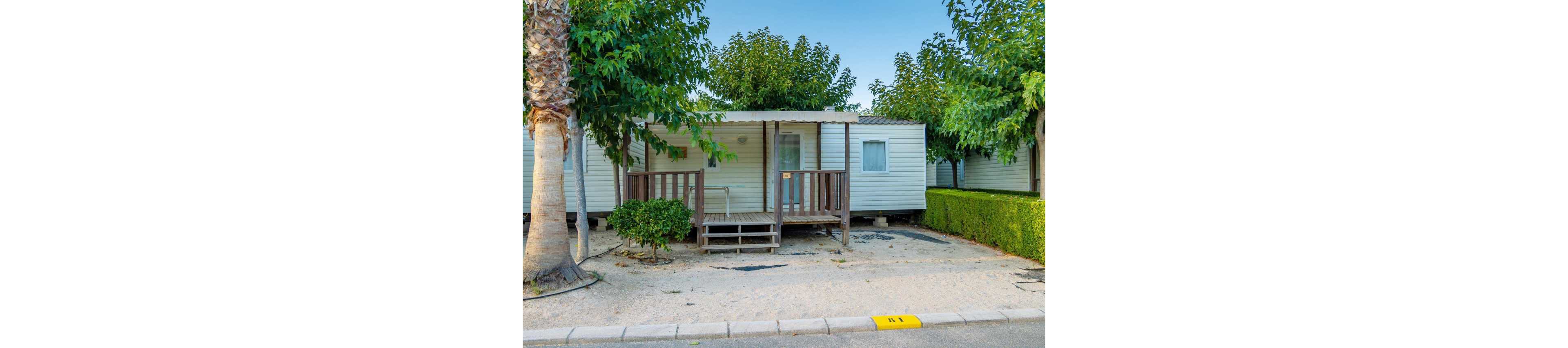 A compact mobile home cabin is parked in a neatly organized campsite. The structure features a small covered porch with steps leading up to the entrance and a white exterior with simple windows. Surrounding the cabin are trimmed hedges, palm trees, and other greenery, creating a relaxed holiday atmosphere. A paved road and curb run in front of the unit, with a yellow marker on the ground indicating the plot number.