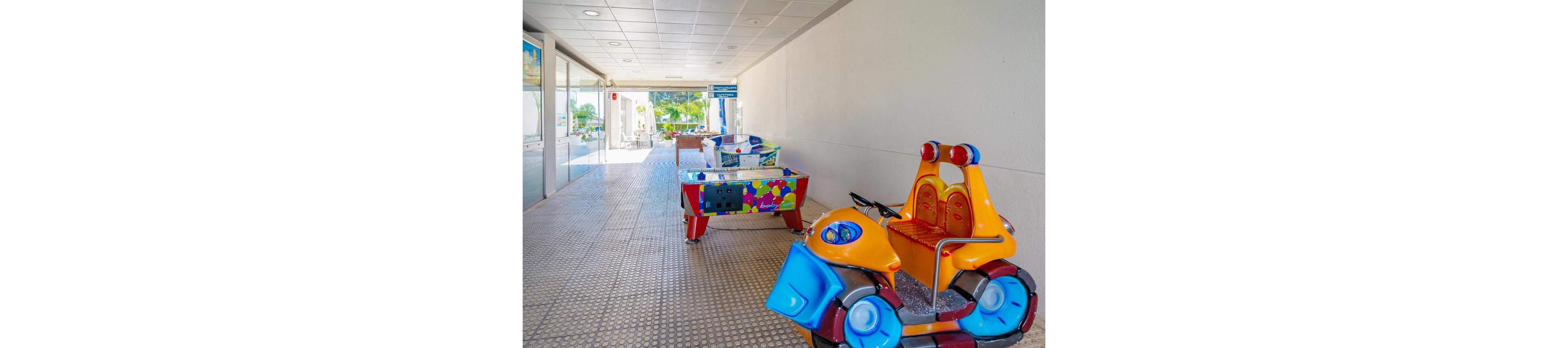A brightly colored children's ride shaped like a motorcycle is placed in a tiled indoor hallway. Further down the corridor, a colorful arcade game table stands near glass doors leading outside. Sunlight filters in from the open end of the passage, revealing greenery and a relaxed leisure setting. The scene suggests a family-friendly entertainment area in a resort or recreational facility.