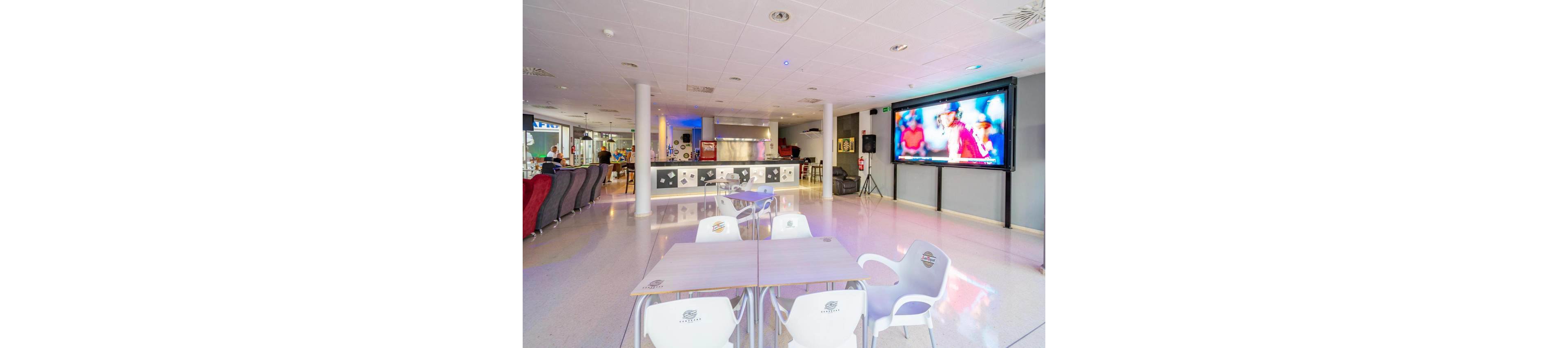 Spacious contemporary lounge area features a bar counter at the back and several tables with white plastic chairs in the foreground. A large flat-screen television on the right wall displays a sports game with players in red uniforms. The room has bright lighting, glossy flooring, and a clean, minimalist design with pops of color from seating and wall decor. The setting suggests a casual social space for watching sports and relaxing.