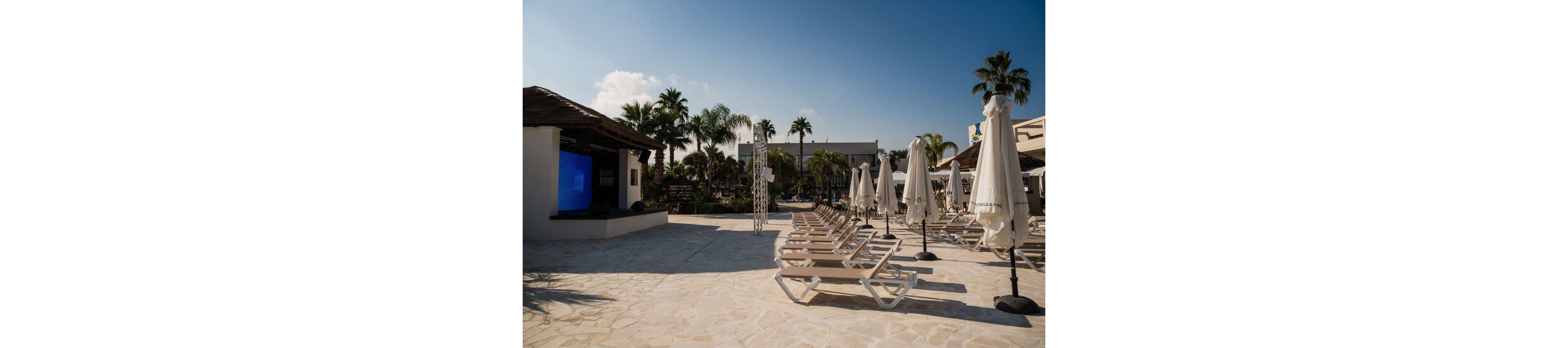 A row of empty sun loungers lines a spacious poolside terrace at a resort. Closed white umbrellas stand between the chairs, casting small shadows on the stone floor. Palm trees and tropical greenery surround the area, with resort buildings visible in the background. The bright daylight and clear sky create a warm, relaxed vacation atmosphere.