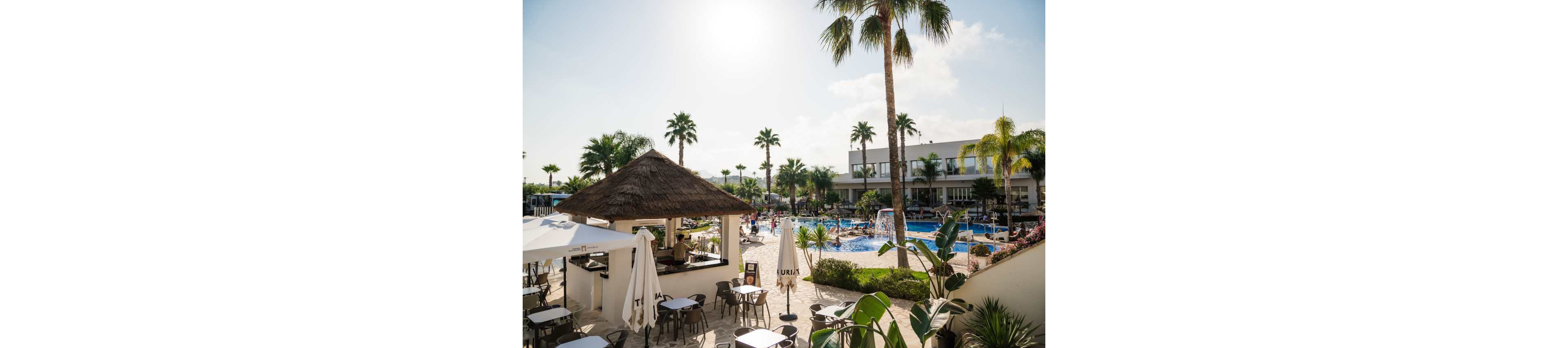 An outdoor resort scene features a thatched-roof poolside bar and shaded terrace seating. Guests relax and swim in a large blue swimming pool surrounded by palm trees and landscaped greenery. White umbrellas and modern buildings frame the leisure area under a bright, partly cloudy sky. The atmosphere suggests a warm, sunny vacation destination focused on relaxation and socializing.