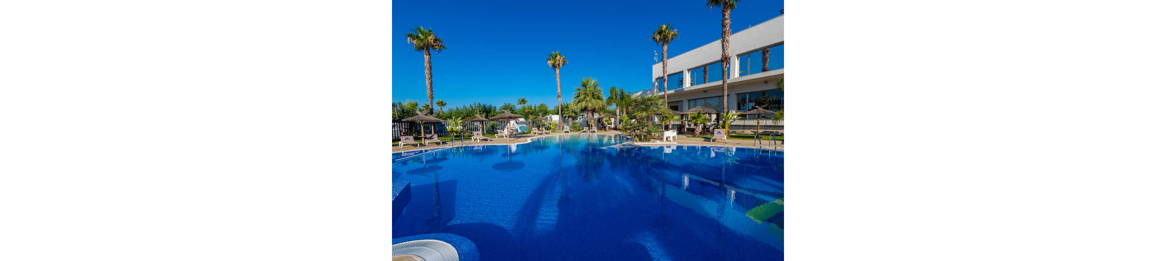 Large outdoor swimming pool reflects a clear blue sky at a modern resort. Tall palm trees and sun loungers with straw parasols surround the water, creating a relaxing vacation atmosphere. A contemporary white building with balconies lines one side of the pool area. The scene is bright and colorful, suggesting a warm, sunny day ideal for leisure and relaxation.