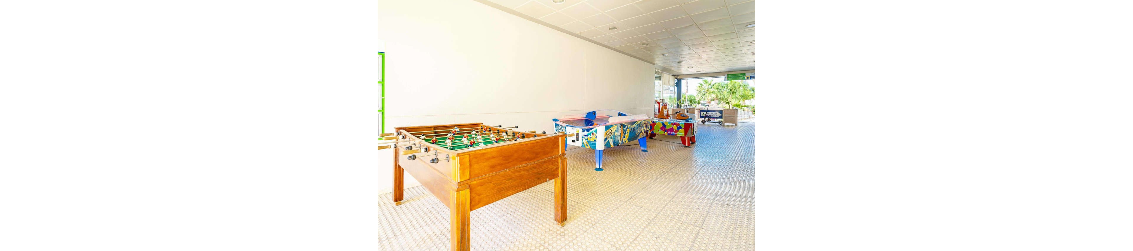A spacious indoor game area features several foosball tables arranged in a row. The nearest table is wooden, while the others have colorful, graphic designs. The room has a tiled floor, white walls, and a grid-patterned ceiling with recessed lighting. Sunlight enters from large openings at the far end, revealing greenery outside and creating a relaxed recreational atmosphere.